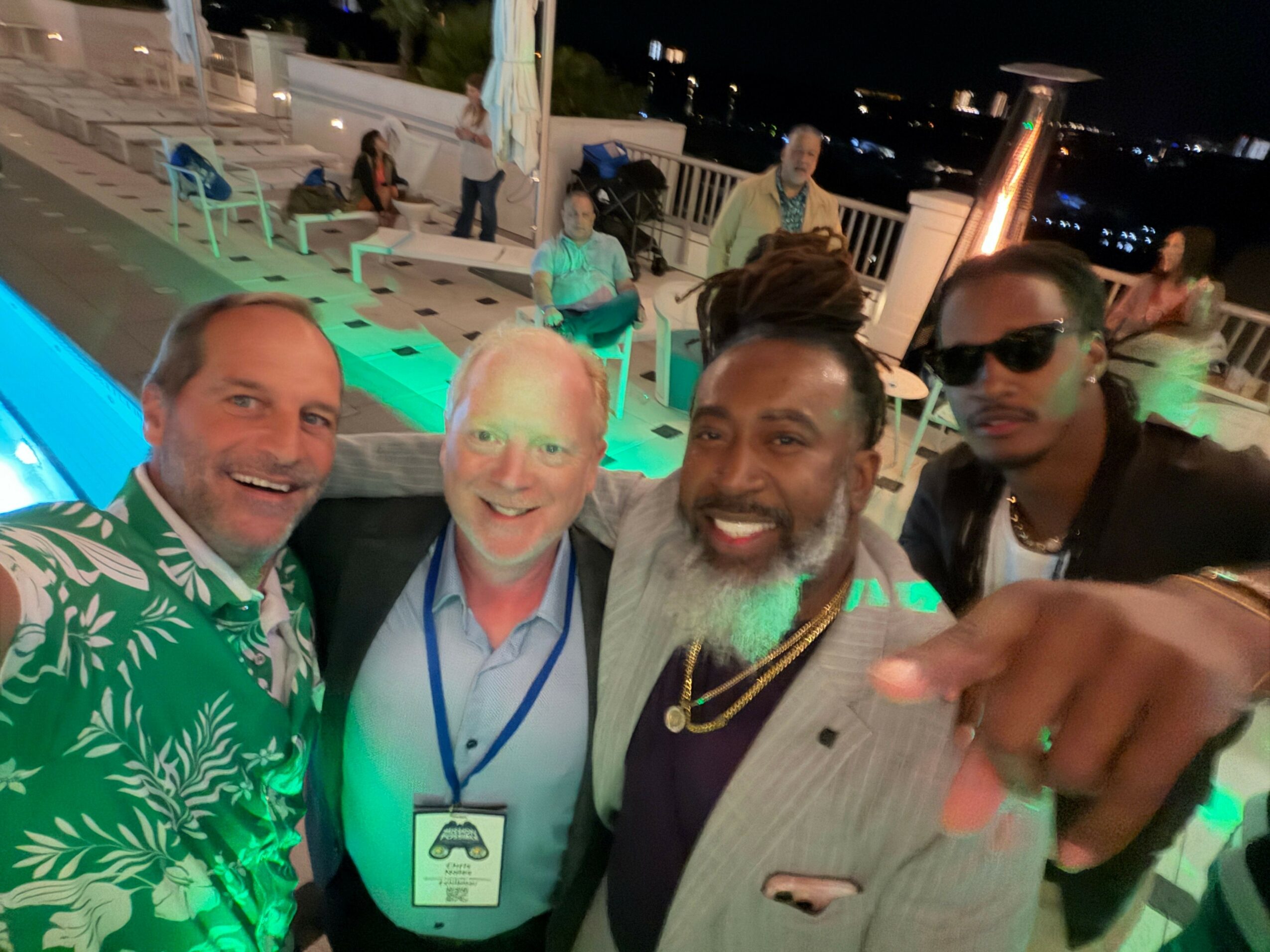 Three people smiling at an outdoor event, with city lights in the background, one wearing a green patterned shirt, one with a name badge, and one with dreadlocks.