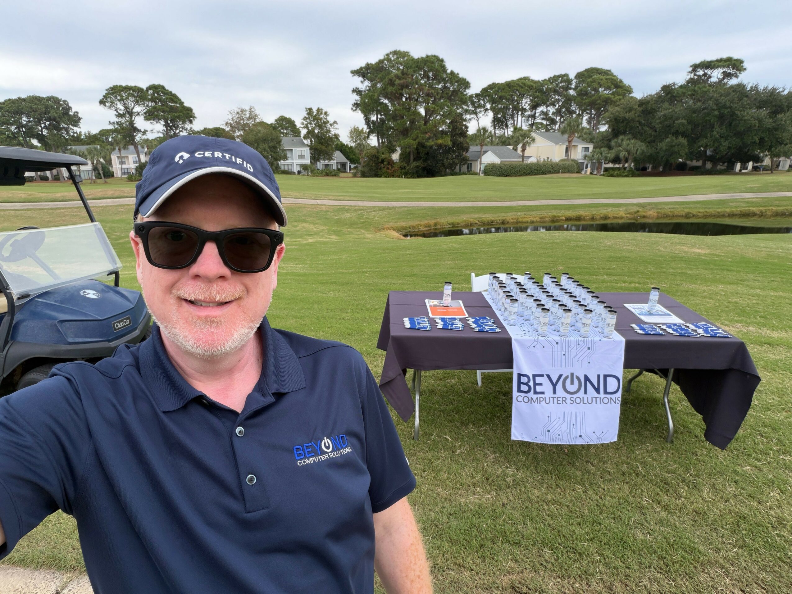 Man wearing sunglasses and a cap stands outdoors near a table with signs and promotional materials, with a grassy area and trees in background.