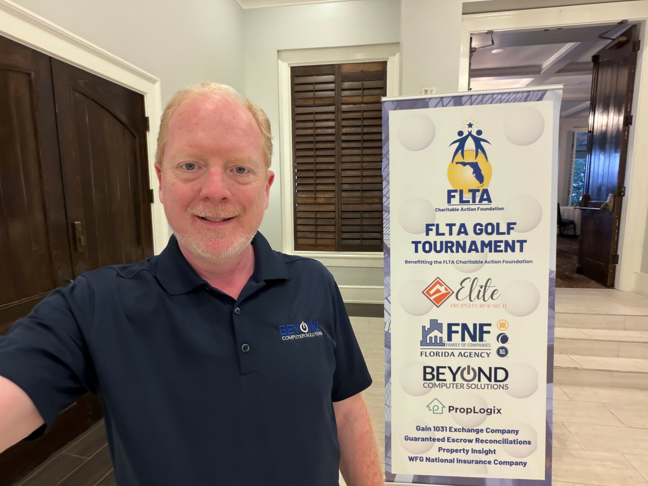 Man with short white hair wearing a dark polo shirt standing indoors near a signboard.