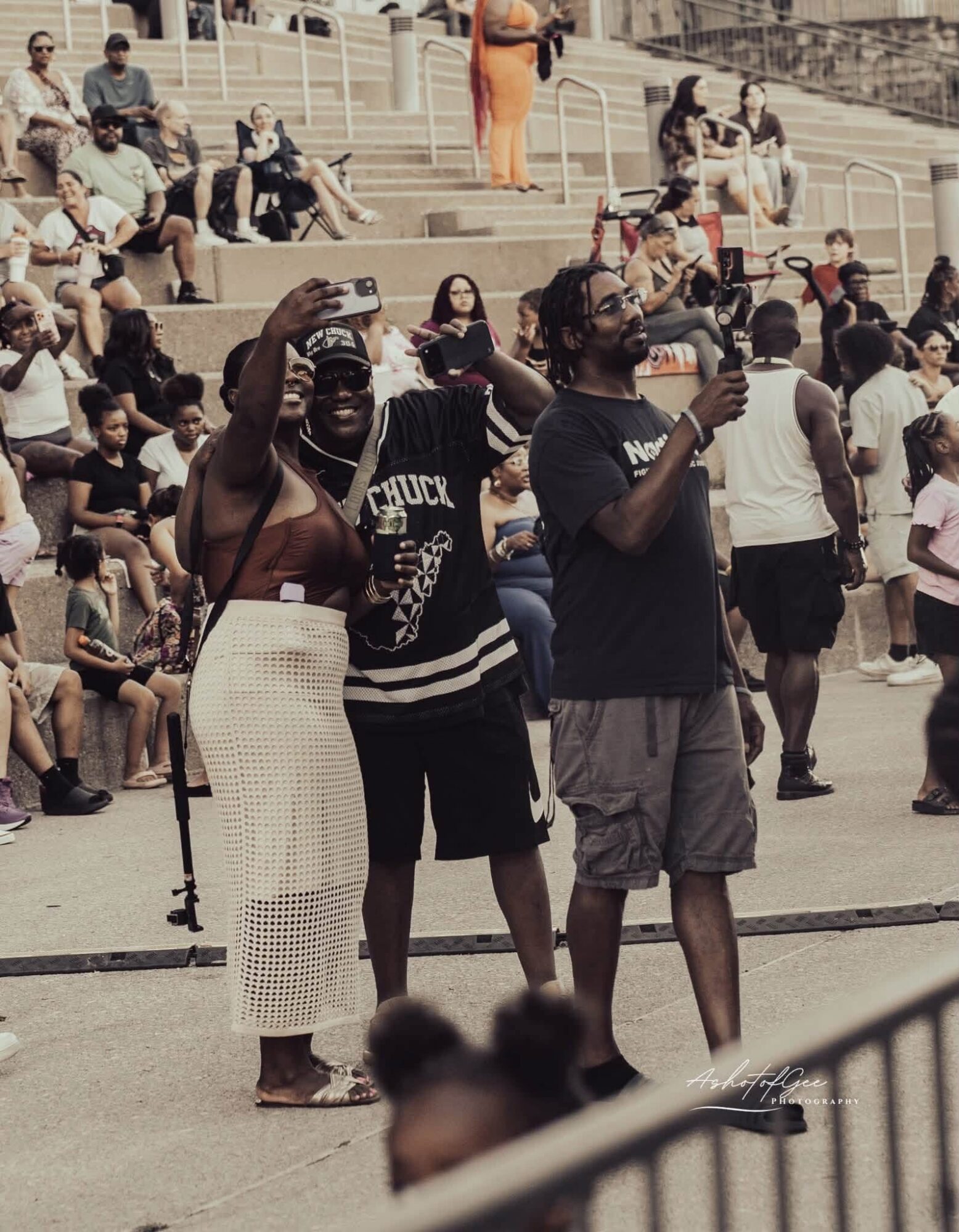 Three people standing outdoors, two taking photos with smartphones, surrounded by seated and standing crowd, some with masks.