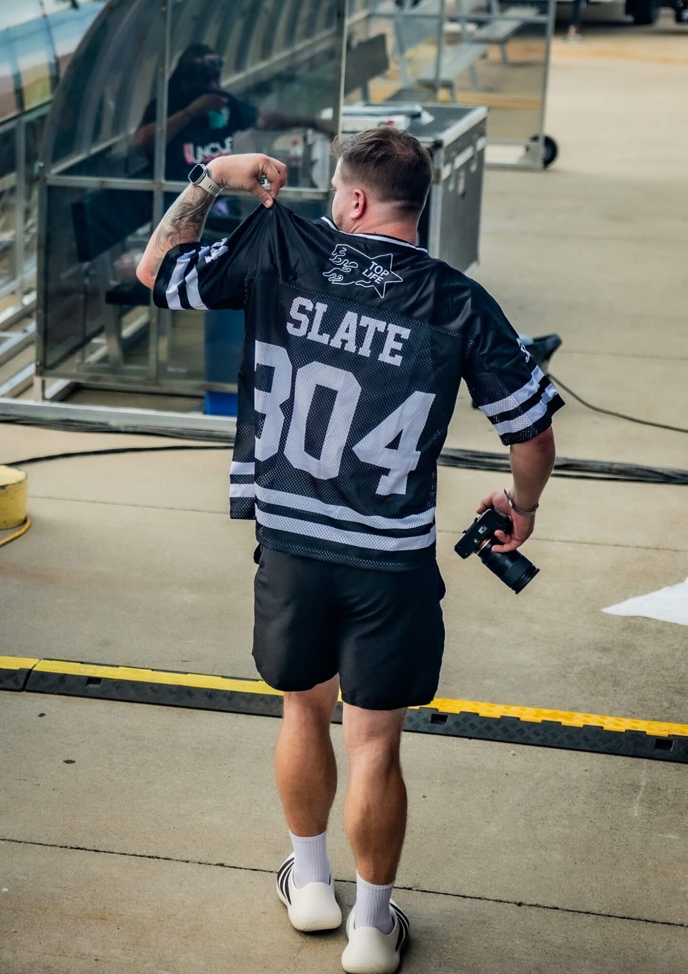 Young man with short hair flexing his arm, holding a camera, wearing a black sports jersey with 'SLATE 304' on the back, shorts, and white shoes, standing outdoors near a glass building.