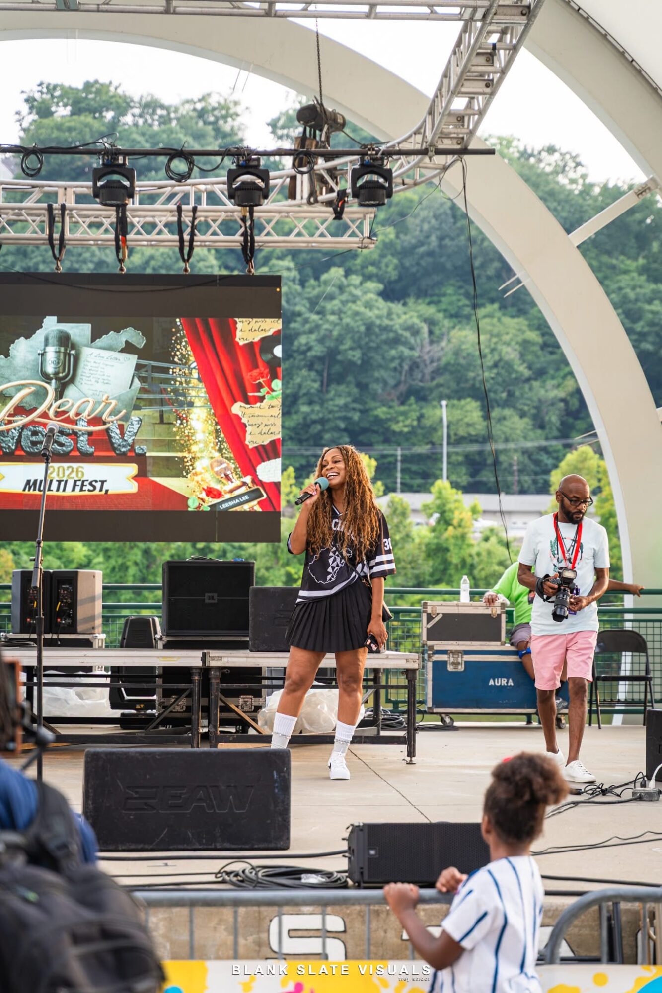 Two performers on stage outdoors, one singing and the other playing guitar, with a large screen and trees in background.