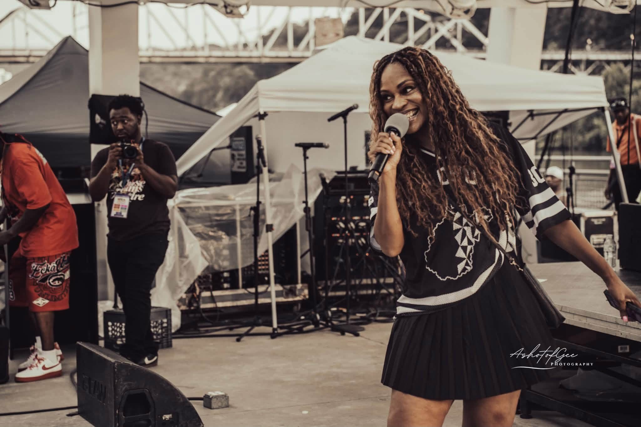 A woman with long curly hair holding a microphone on stage at an outdoor event, with people and tents in the background.