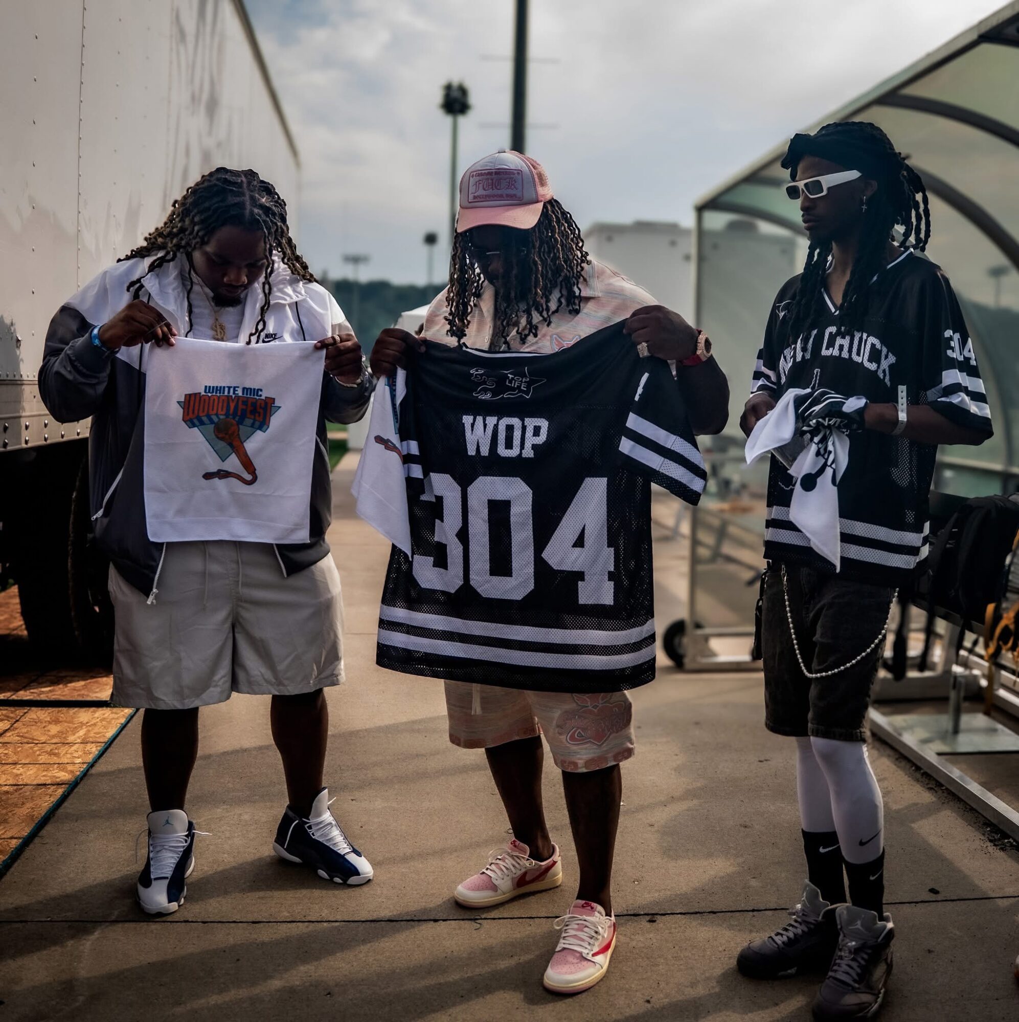 Three people standing outdoors, holding sports jerseys, with a cloudy sky and structures in the background.