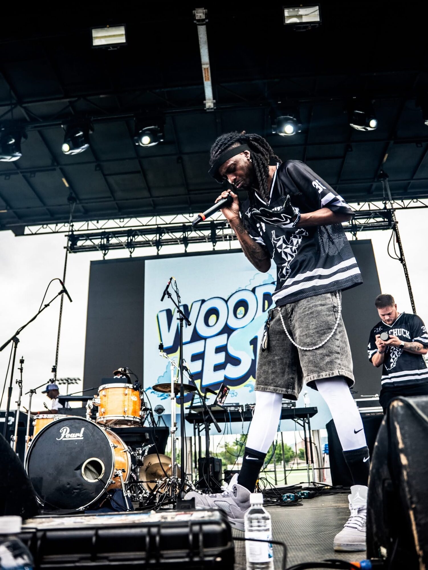 Performer with dreadlocks singing into microphone on stage with band equipment and a large screen behind, outdoors.