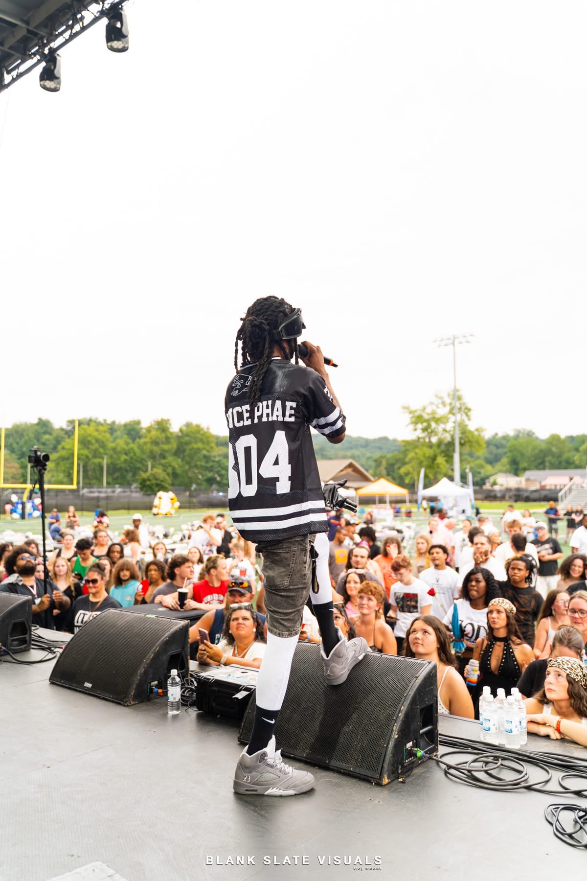 Performer on stage with a microphone, wearing a black and white jacket, facing a crowd outdoors during daytime.