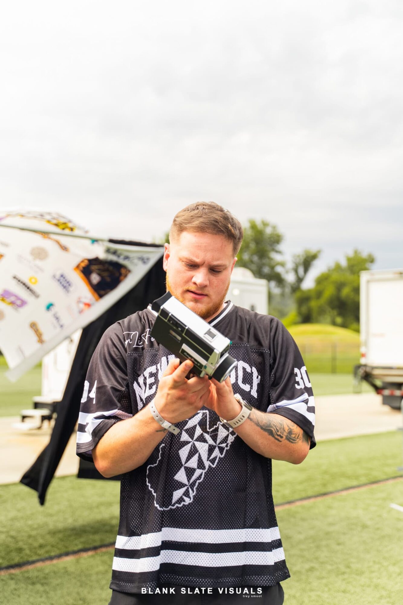Man with tattoos wearing a black sports jersey looks at a camera outdoors on cloudy day.