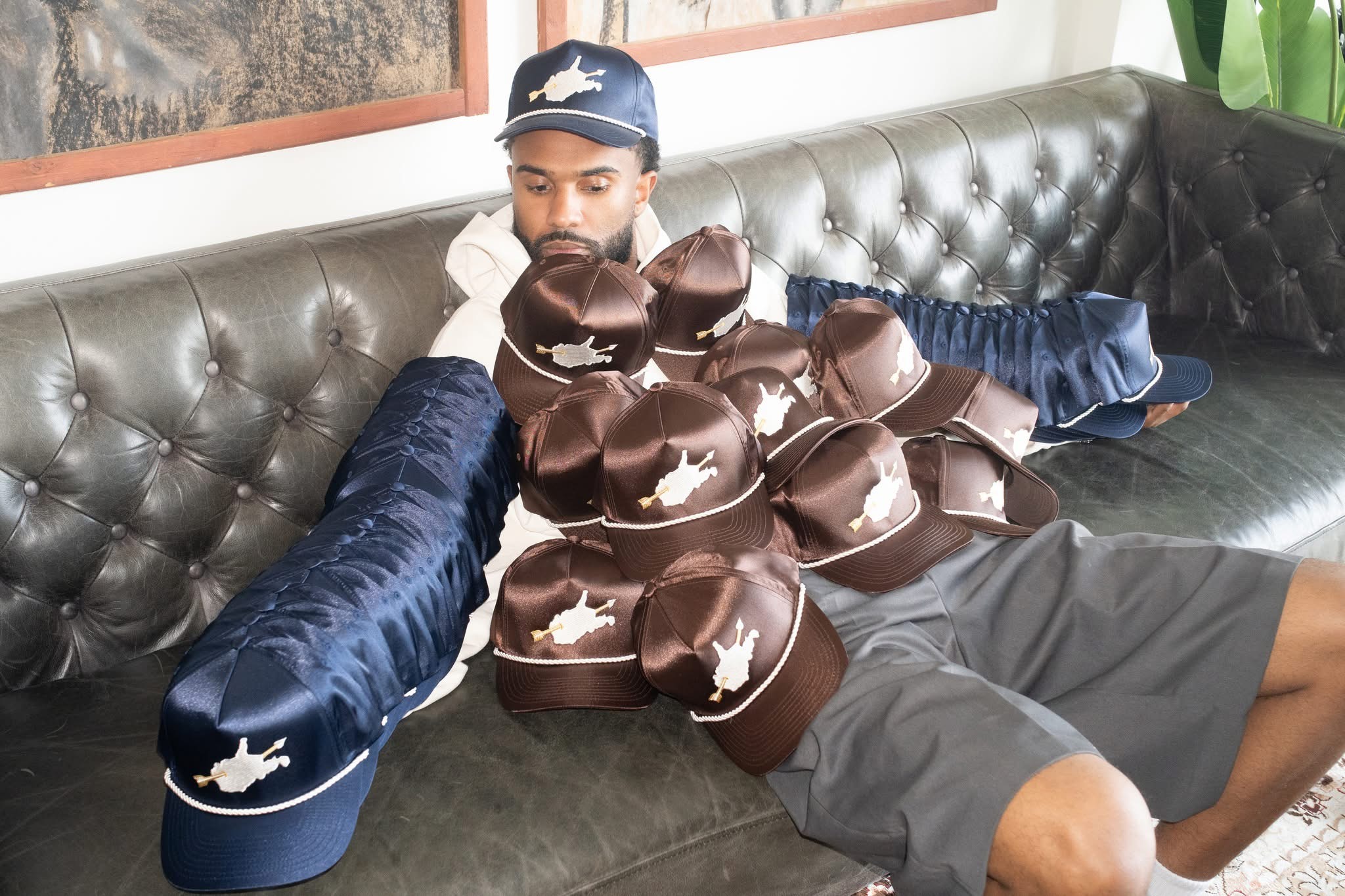 Person lying on a black leather couch surrounded by numerous chocolate cupcakes with white animal-shaped decorations, wearing a cap and casual clothing.