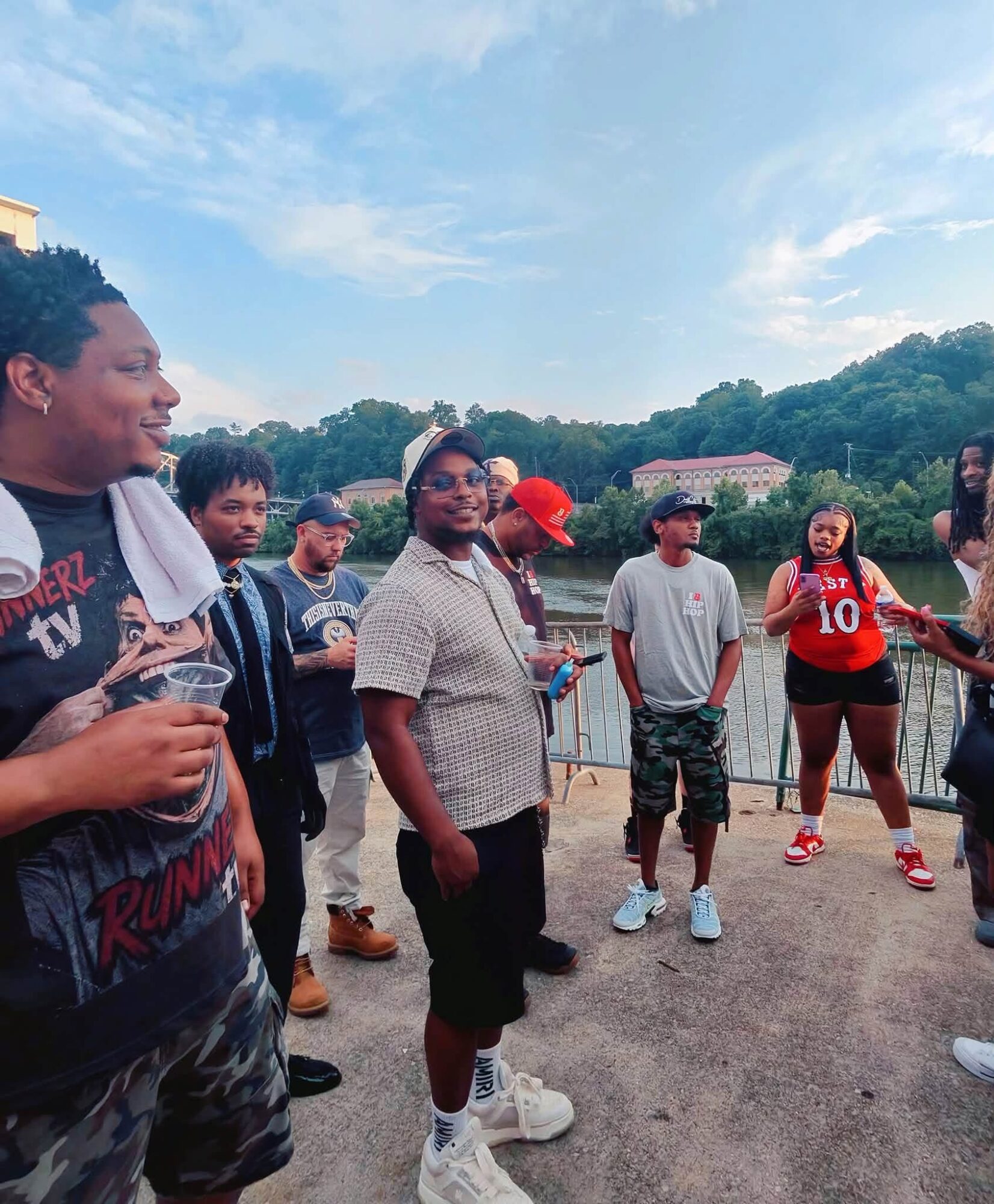 Group of people outdoors, some talking and holding drinks, with trees and buildings in the background.