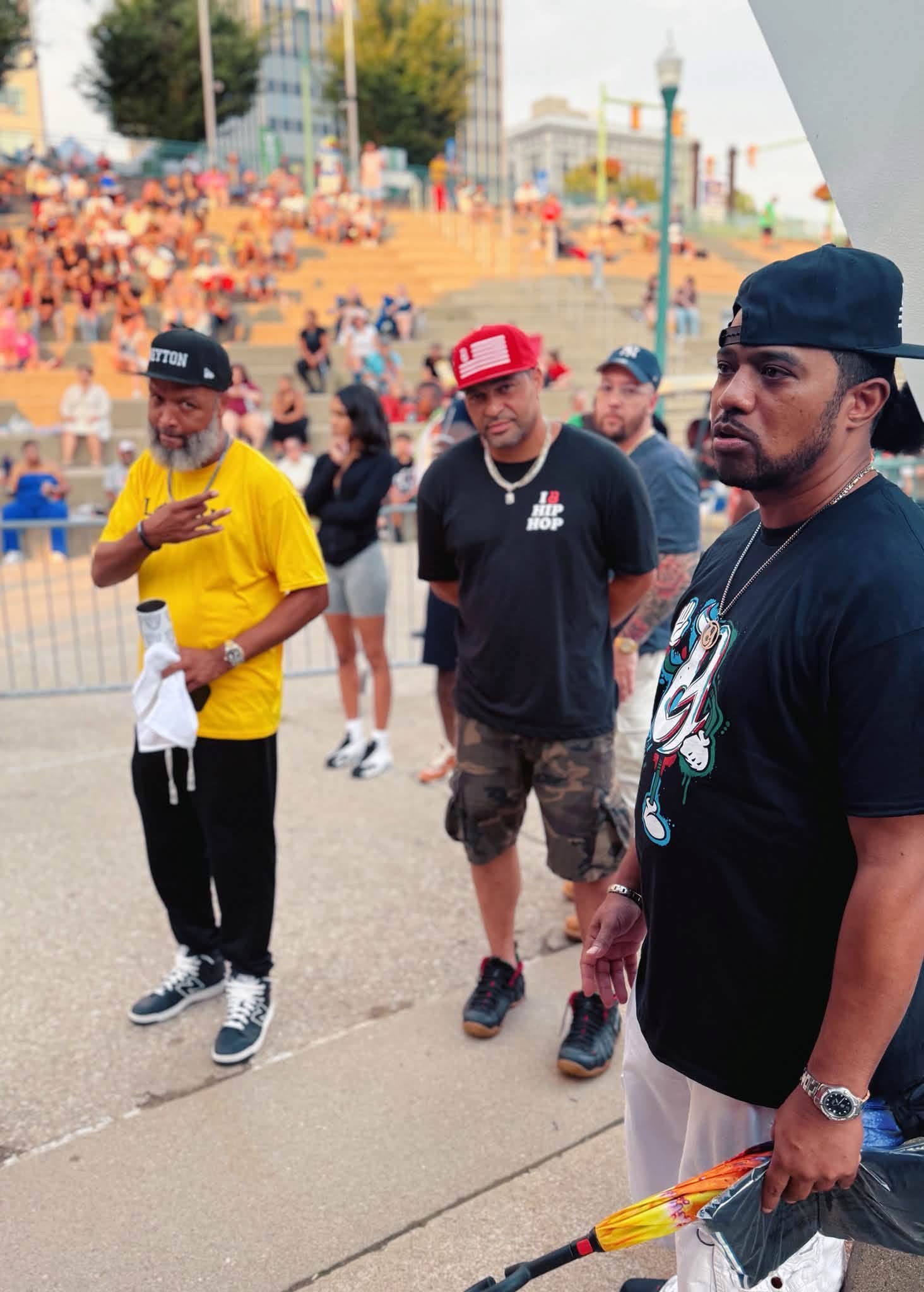 Three men standing outdoors in a crowd, one in a yellow shirt, one in a black shirt with a red cap, and one in a black shirt with a black cap, holding a colorful object, with a large audience in the background.
