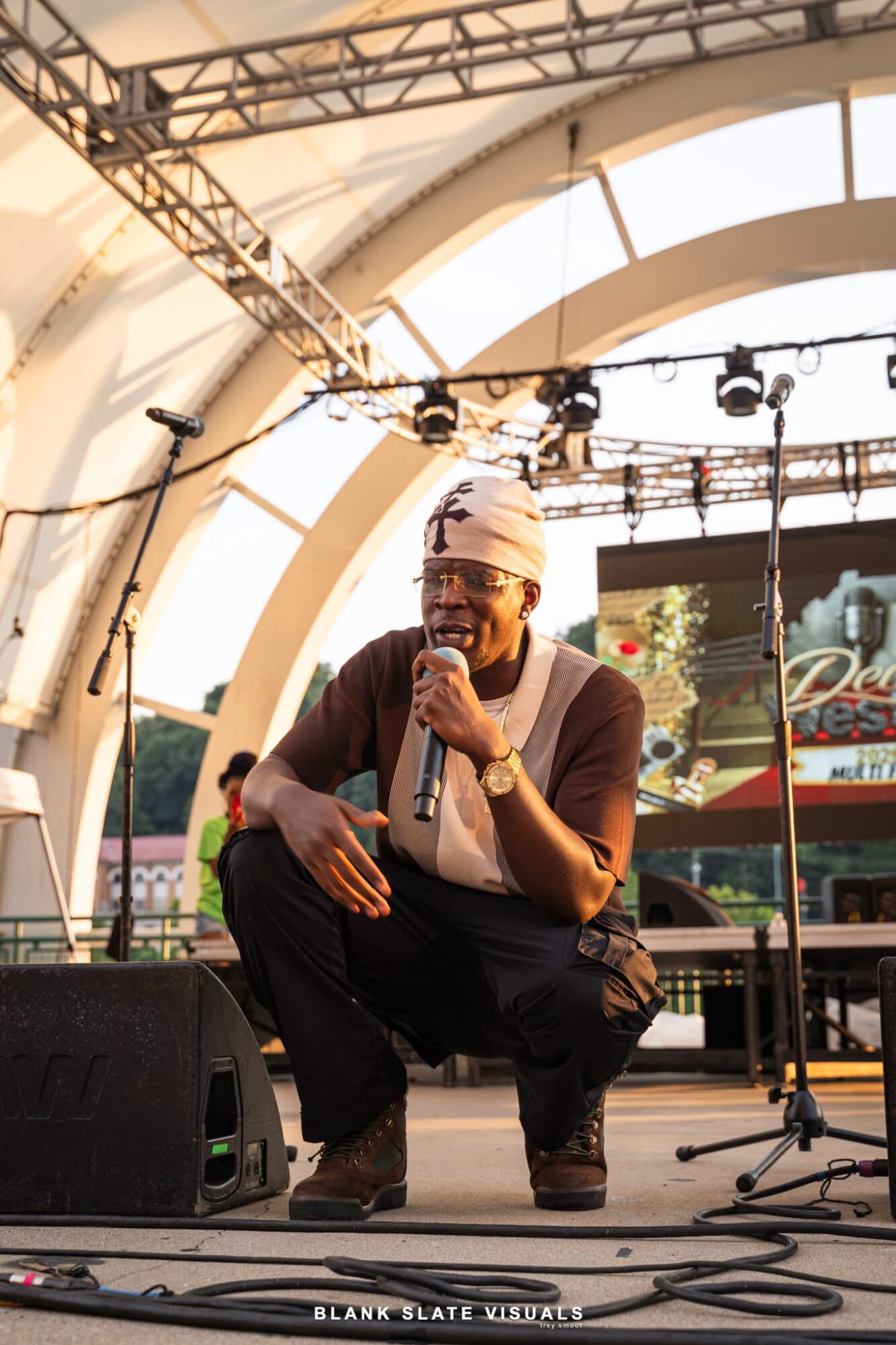 Person crouching on stage with microphone, wearing glasses and a headwrap, under a large arched structure, stage equipment visible.
