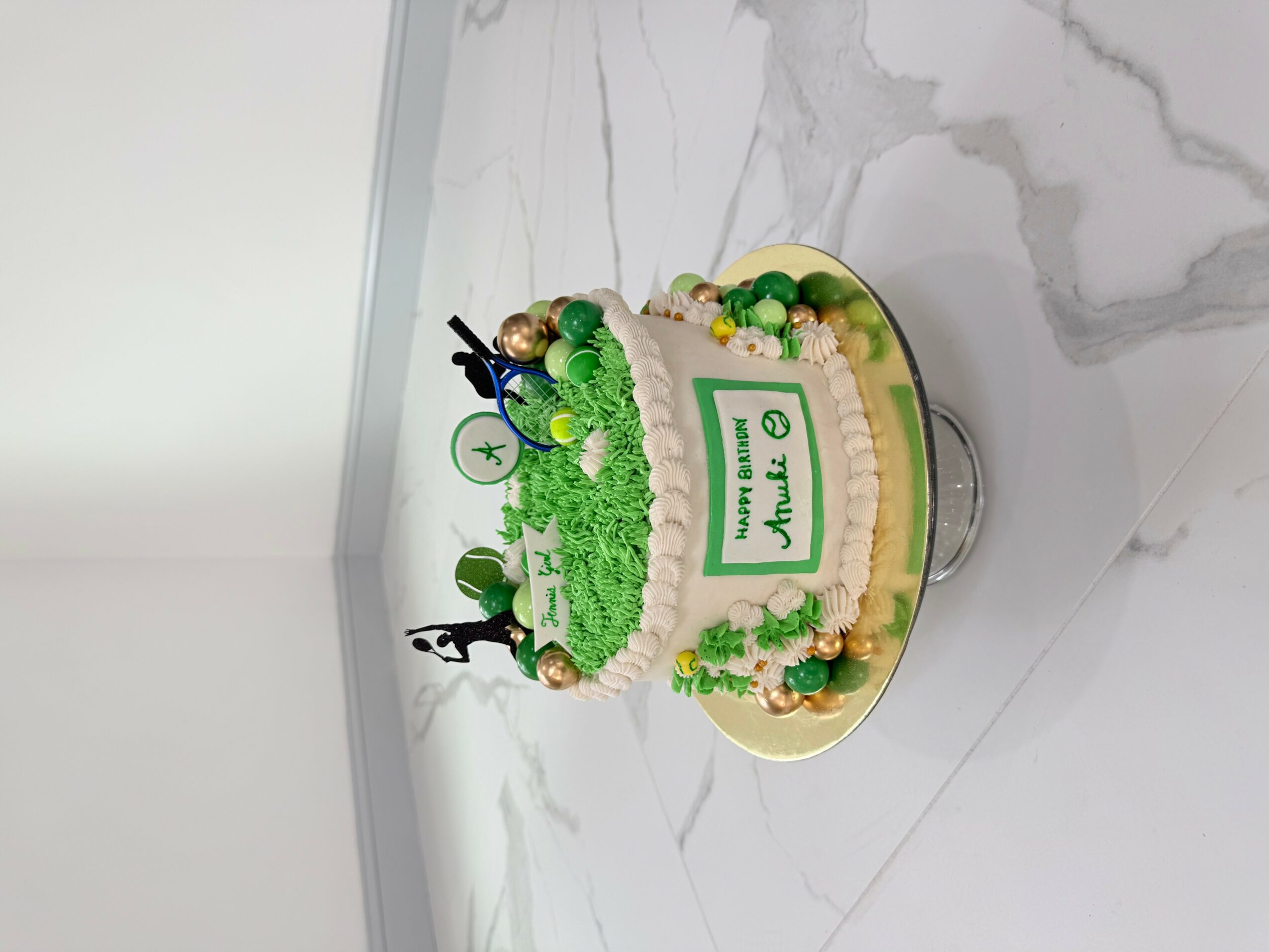 Round birthday cake with green and white decorations, topped with small figures and a 'Thank You' sign, on a marble surface.