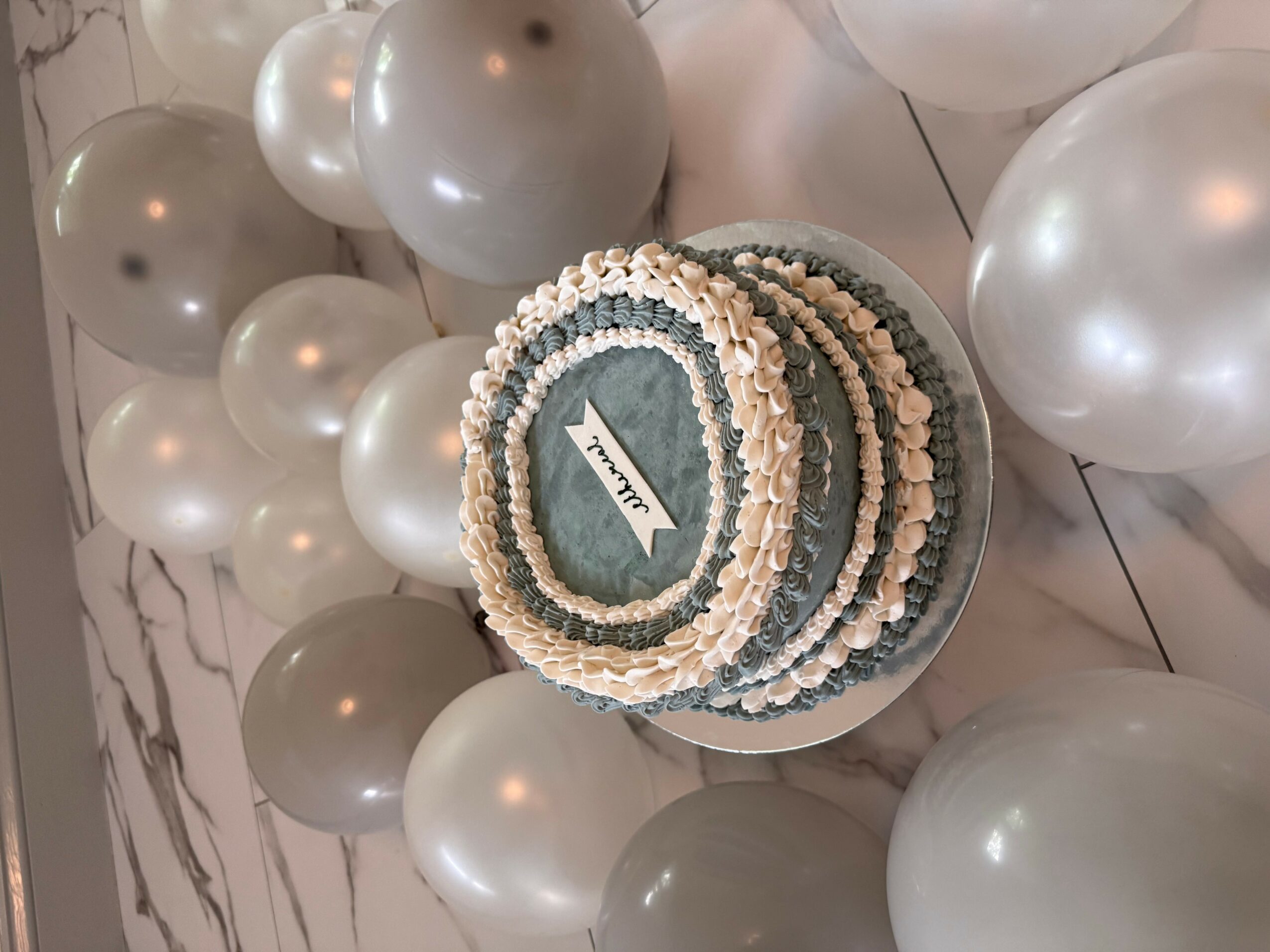 Round cake with white and blue icing, surrounded by silver and white balloons, on a marble surface.