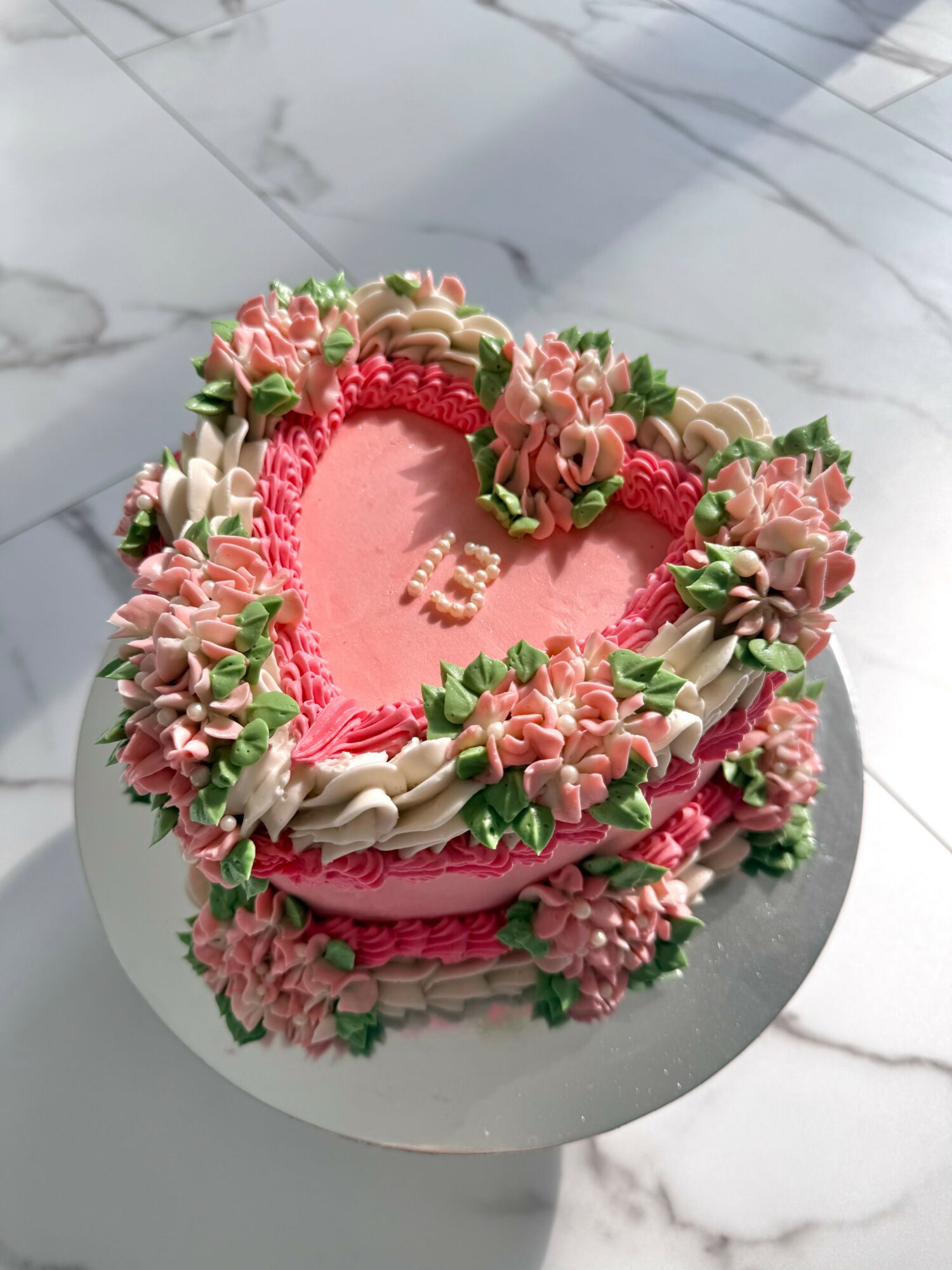 Heart-shaped cake decorated with pink and white flowers, green leaves, and a pink border, on a white marble surface.