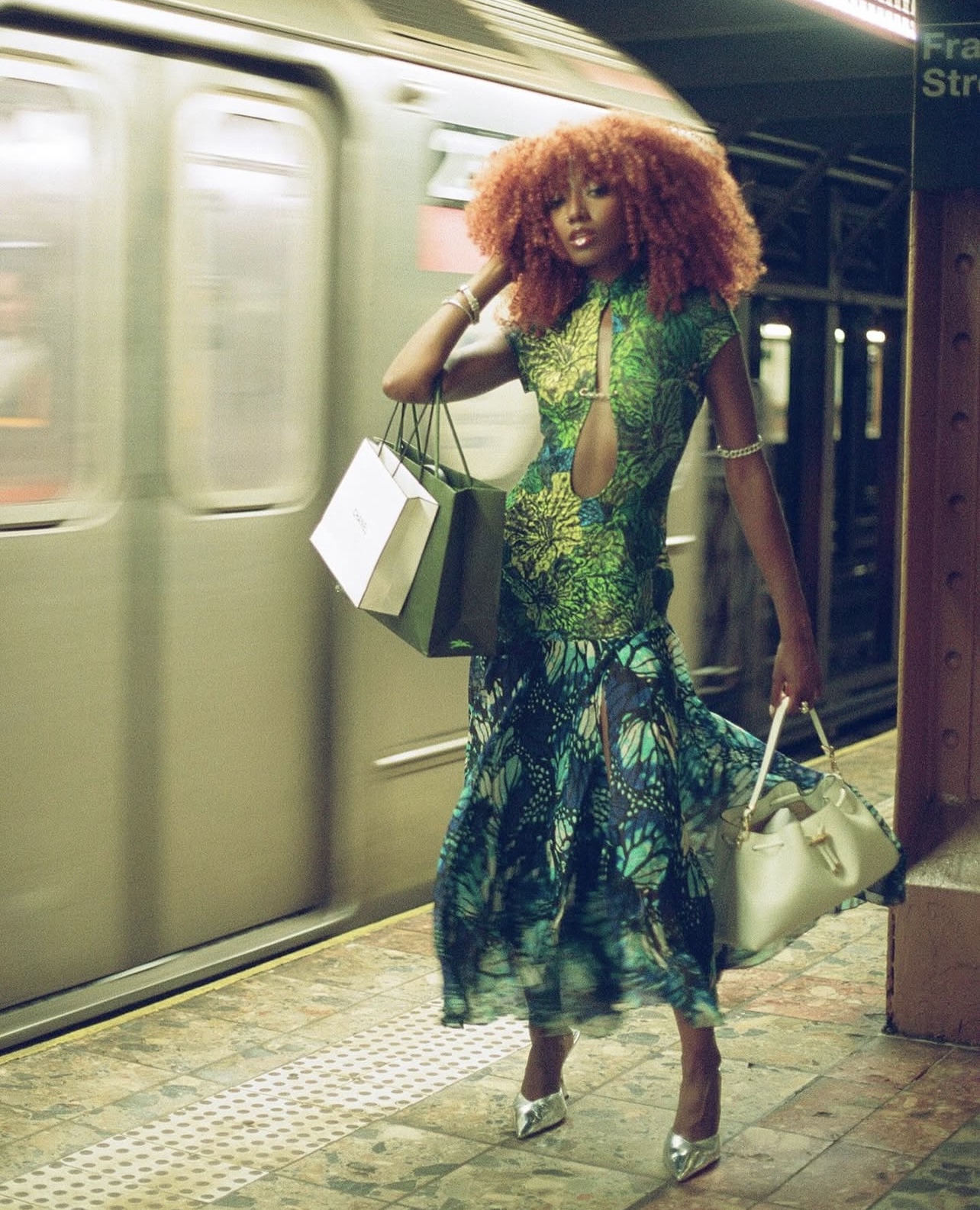 Woman with curly hair in a colorful dress standing on subway platform holding shopping bags and a purse.