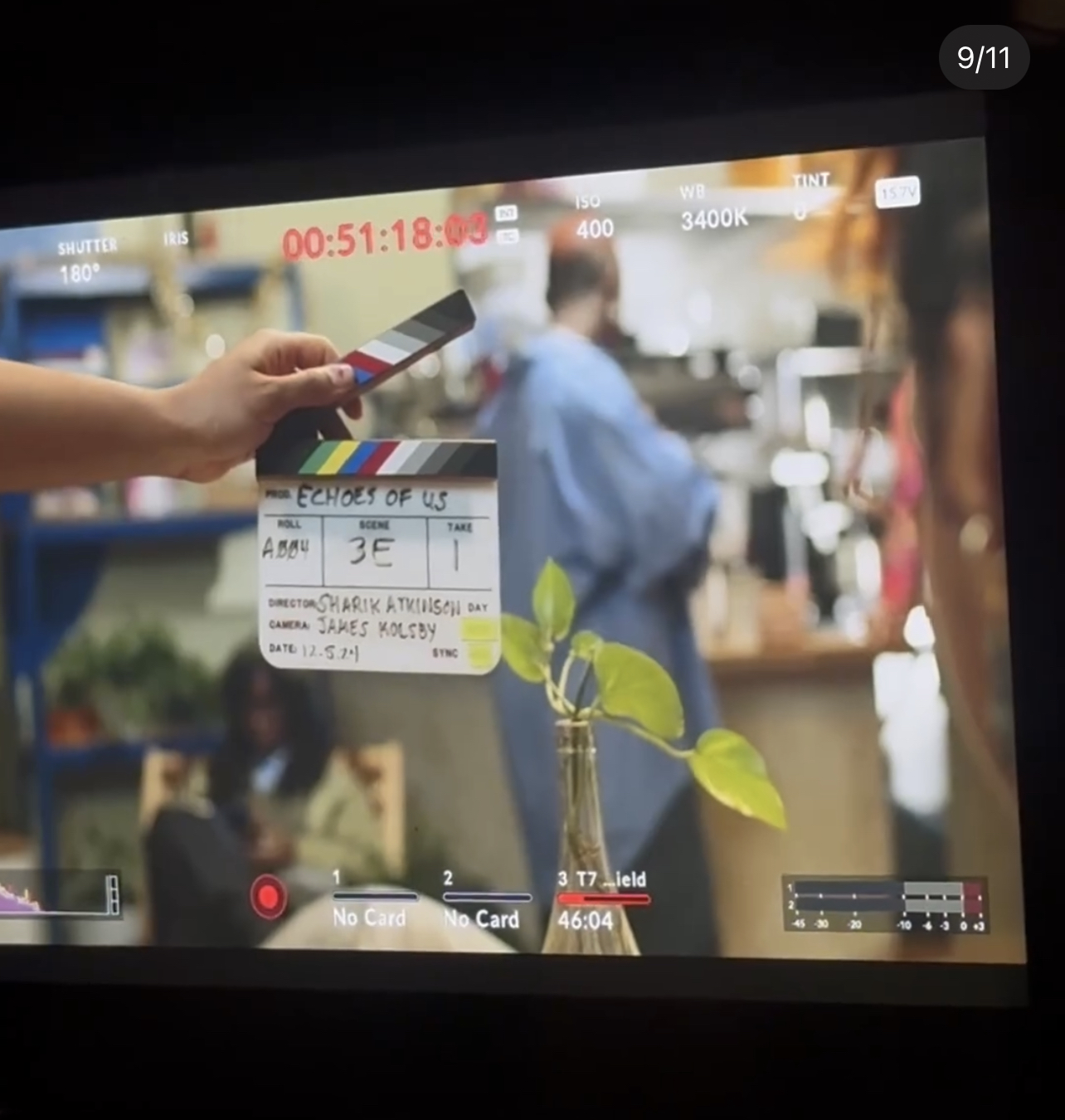 Close-up of a hand holding a clapperboard in a busy indoor setting with people in the background.