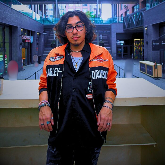 Young man with glasses and curly hair stands indoors, wearing an orange Harley-Davidson jacket over a black shirt, with accessories.