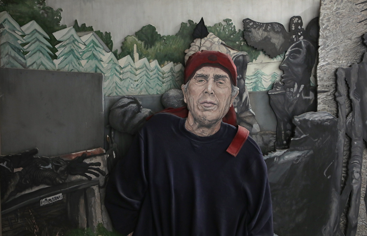 Older man with a red hat and dark clothing standing in front of a mural with trees and faces, with a black dog nearby.