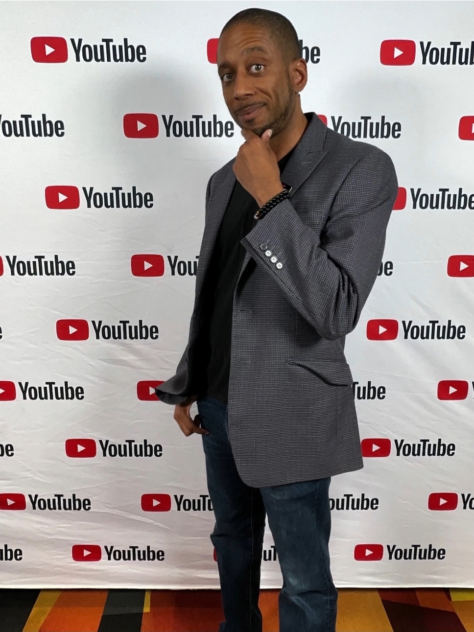 Man in gray blazer and jeans standing in front of a YouTube backdrop, hand on chin, looking at camera.