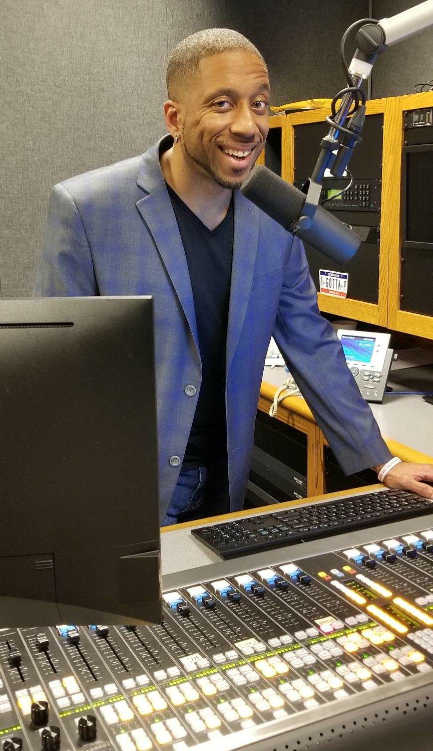 Man in a blue blazer smiling at a radio station with equipment and microphone.