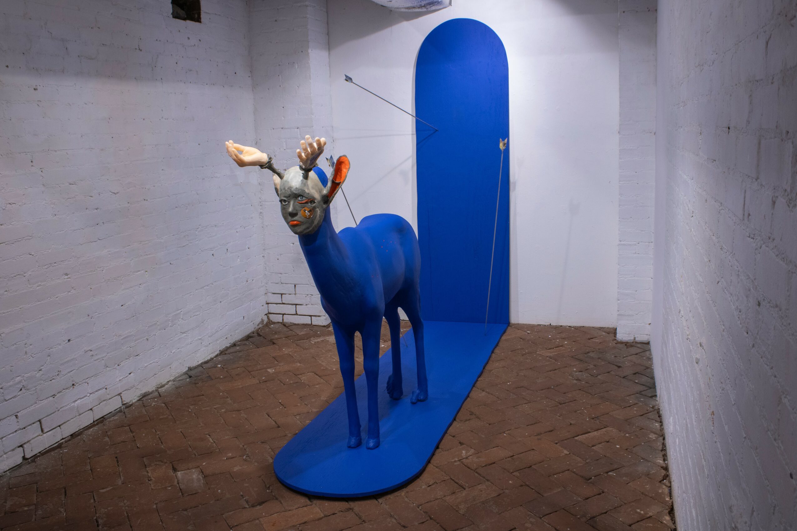 Blue sculpture of a deer with antlers, standing on a blue platform in a white-walled room.