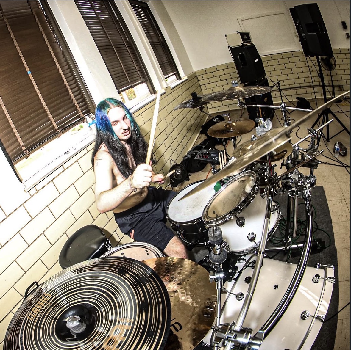 Person with long dark hair playing drums in a room with windows and musical equipment.