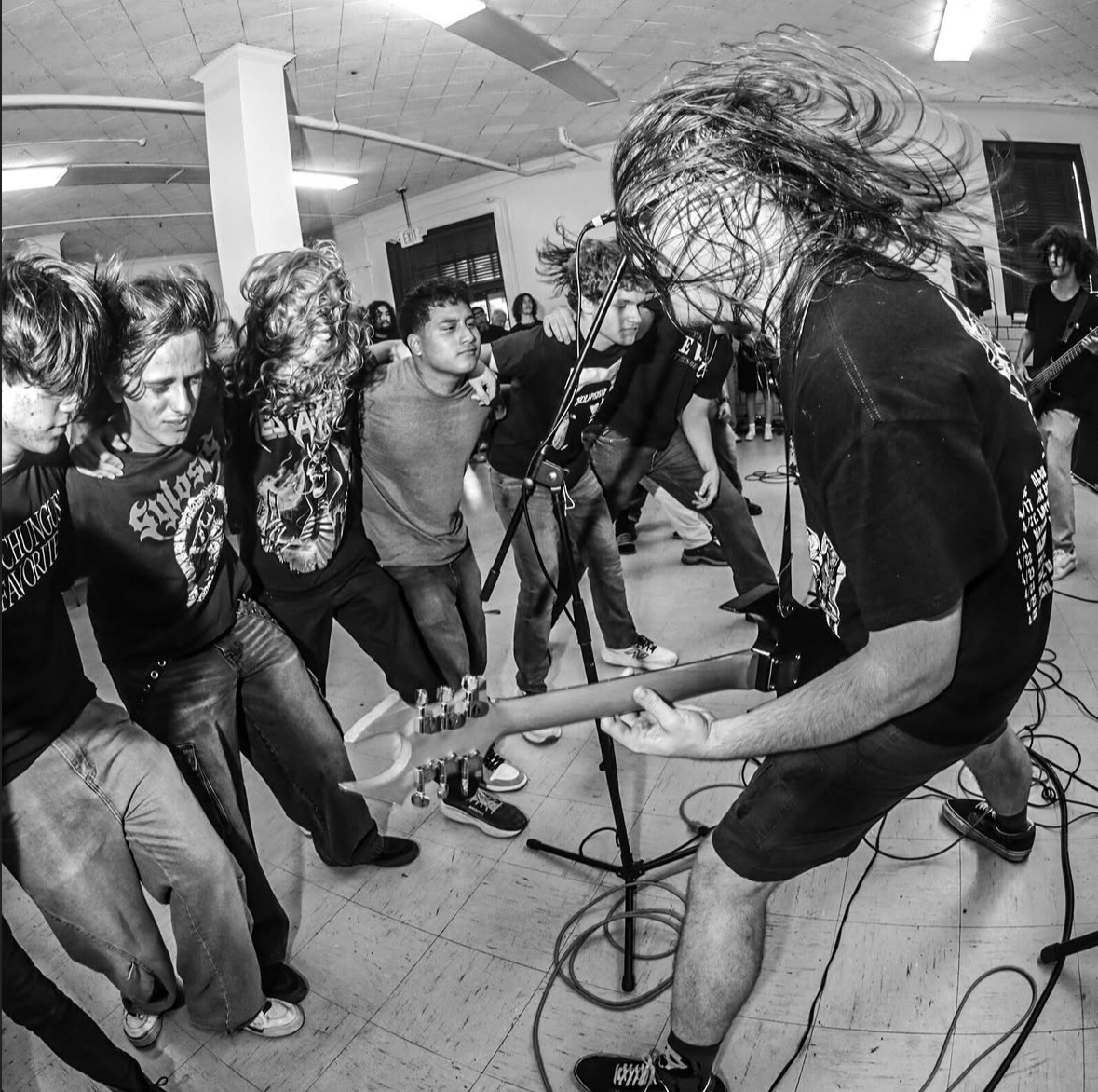 Musician with long hair playing guitar on stage, audience watching closely in a room with ceiling lights.