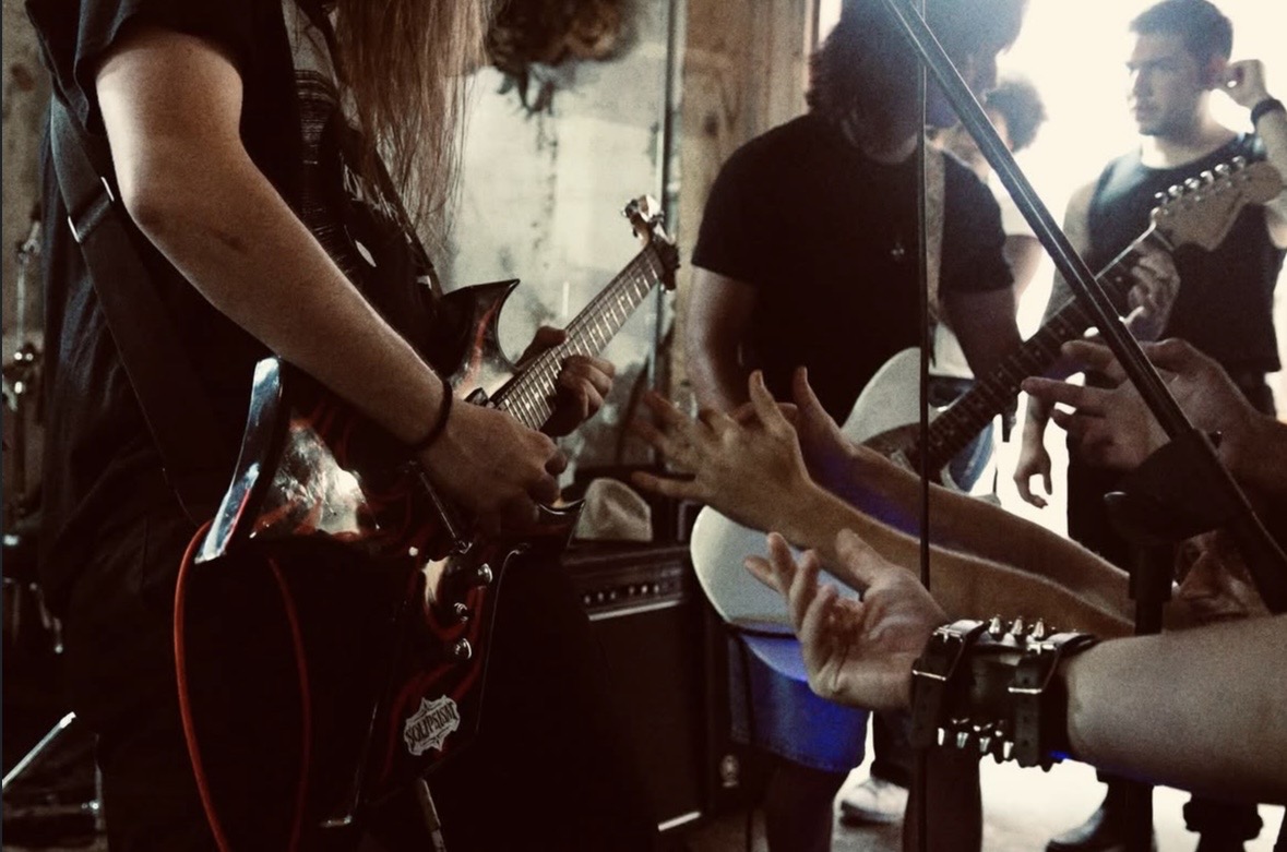Group of musicians playing guitars and drums in a room with natural light, hands and instruments visible.