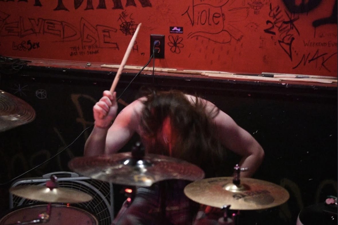 Person with long hair head down, holding drumsticks, surrounded by drums and graffiti-covered wall.