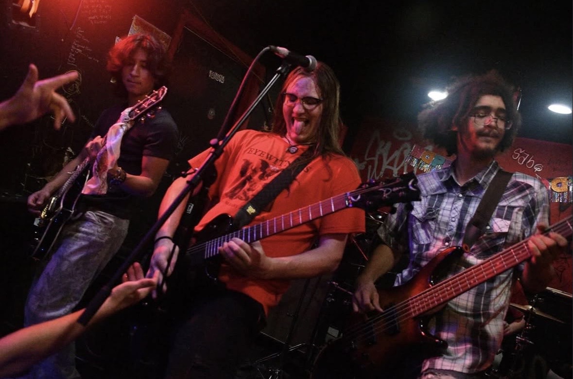 Three musicians performing on stage, one playing guitar, one singing and playing guitar, and one playing bass guitar, with stage lighting.