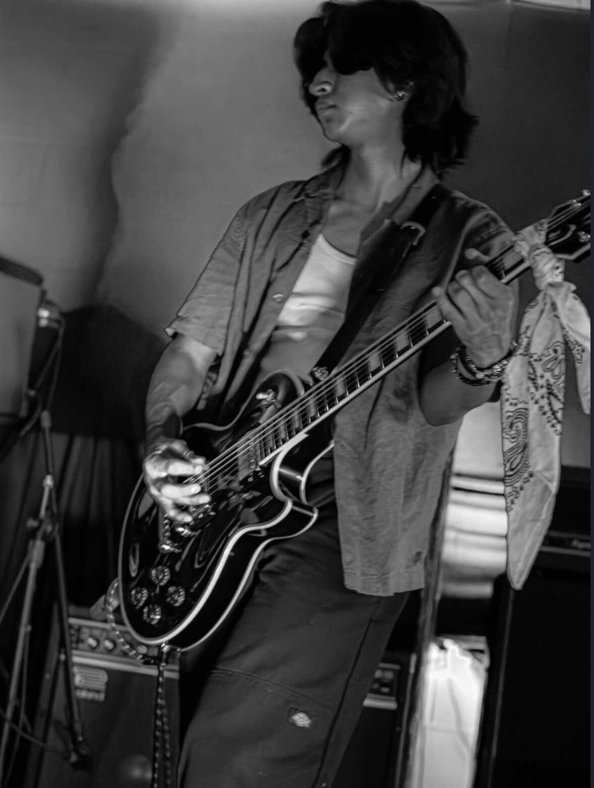 Person playing an electric guitar, smiling, with dark hair, in a room with musical equipment, in black and white.