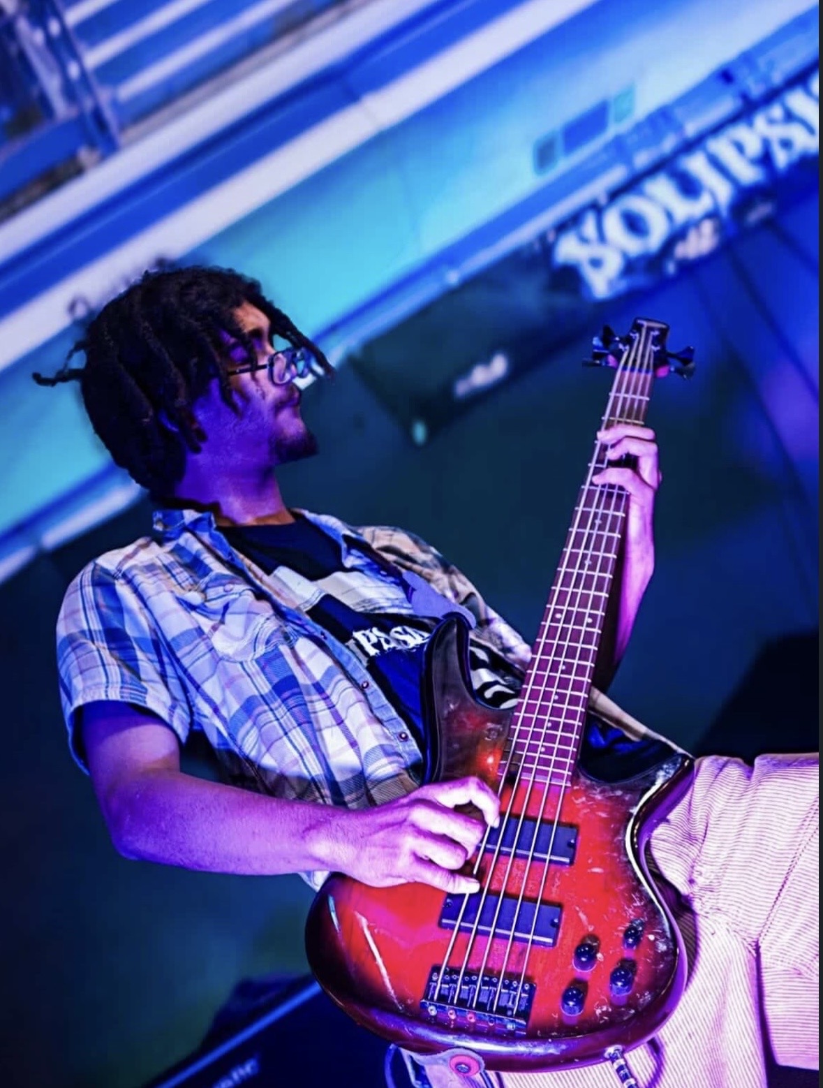 Person playing a red electric bass guitar on stage with blue lighting, wearing glasses and a plaid shirt.