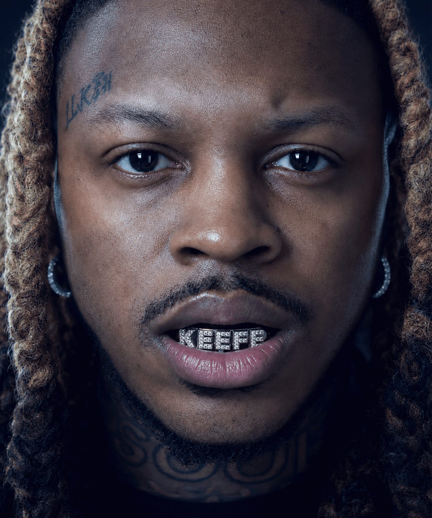 Close-up of a person with dreadlocks, facial tattoos, earrings, and a grill on their teeth, looking directly at the camera.