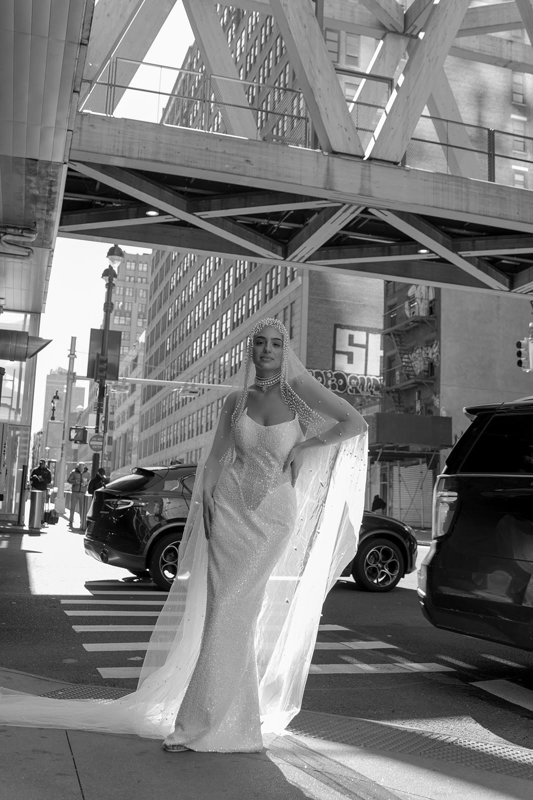 Woman in a long dress walking on city street with cars and buildings, sunlight casting shadows.