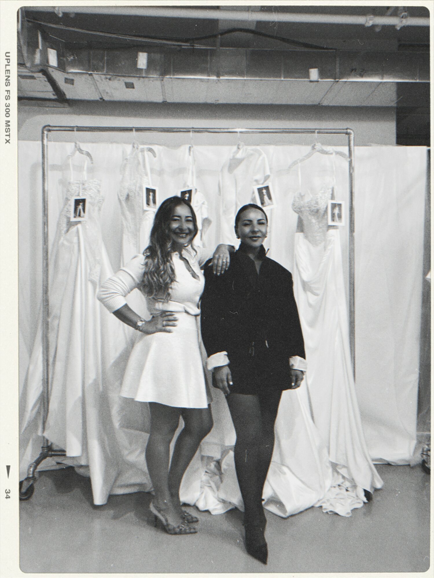 Two women stand together in front of a backdrop with hanging dresses, smiling, one with hand on hip, the other with arm around.
