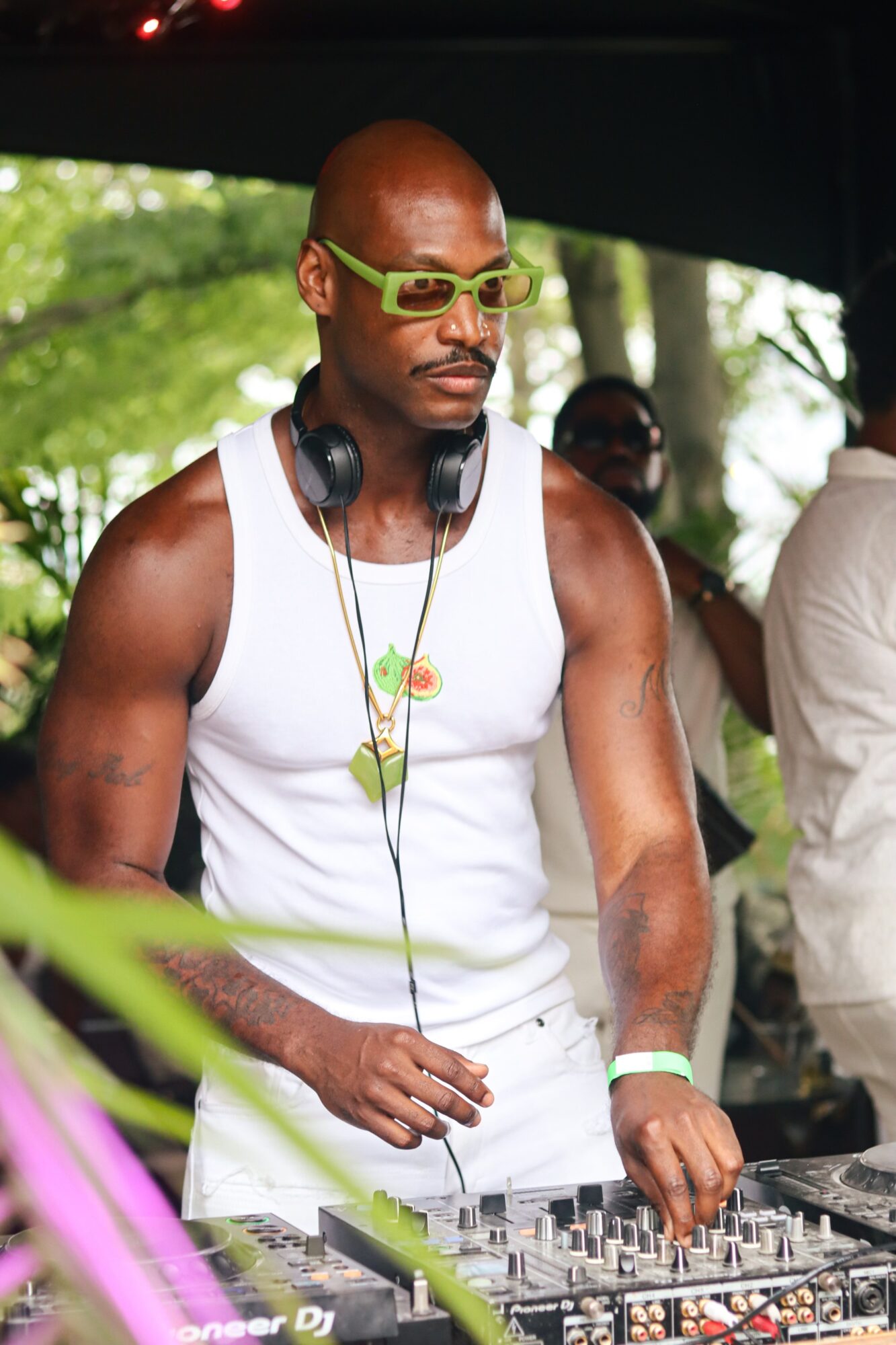 Man with sunglasses and headphones stands outdoors with trees in background, wearing a white tank top and a flower necklace.
