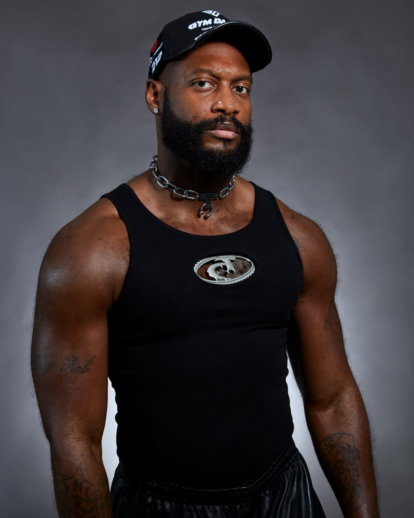 Man with a beard wearing a black tank top, cap, and chain, standing against a gray background.