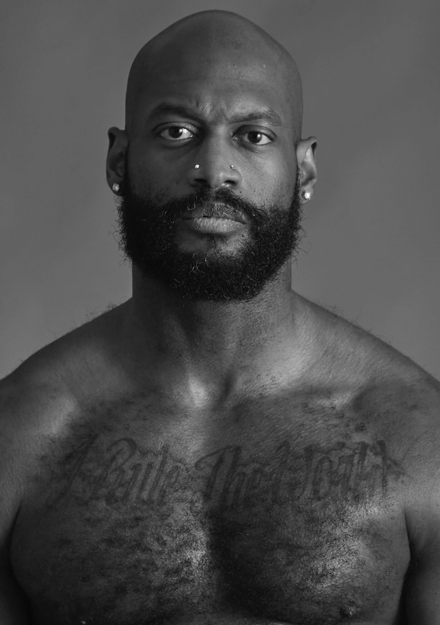 Black and white portrait of a shirtless man with a beard, earrings, and tattoos on his chest, looking directly at the camera.