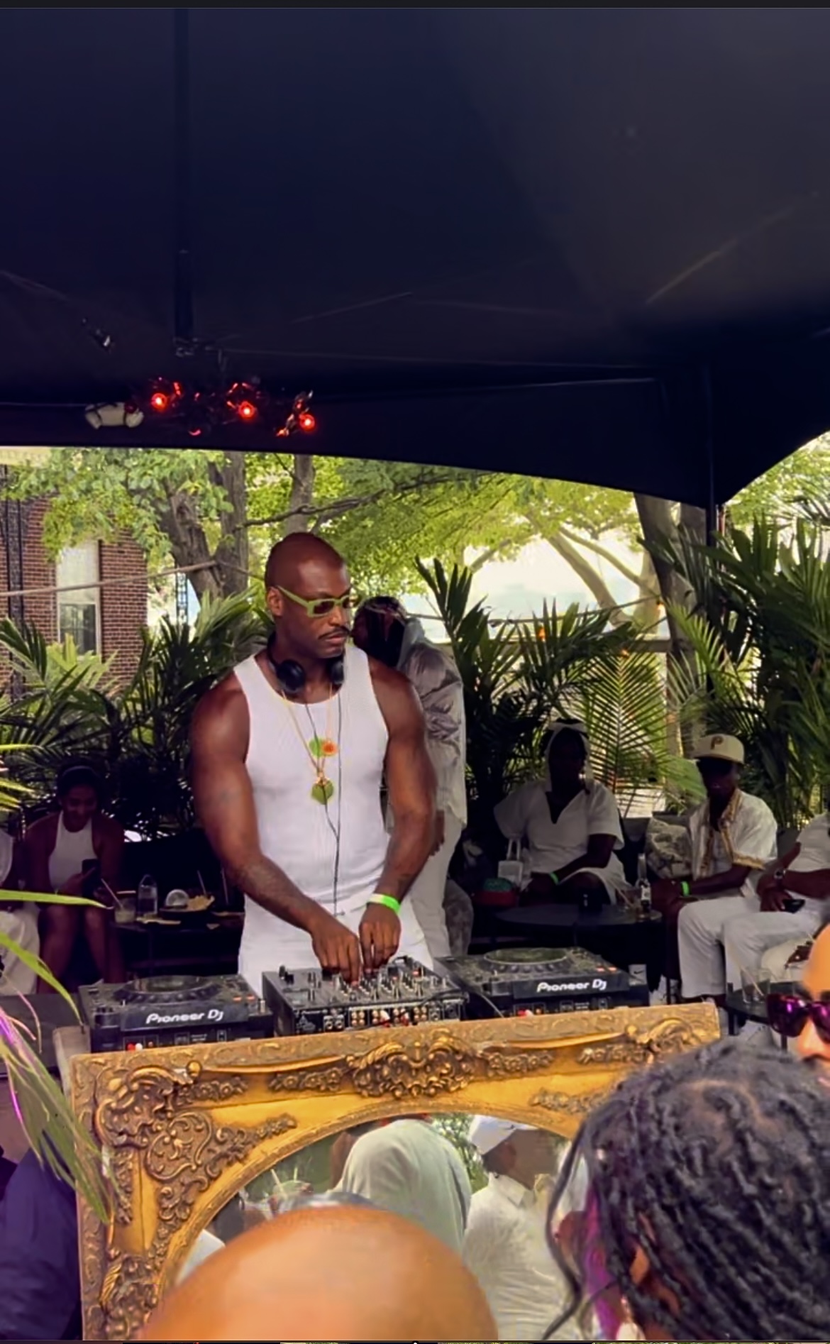 DJ wearing sunglasses and a white tank top, performing at an outdoor event with people and greenery in background.