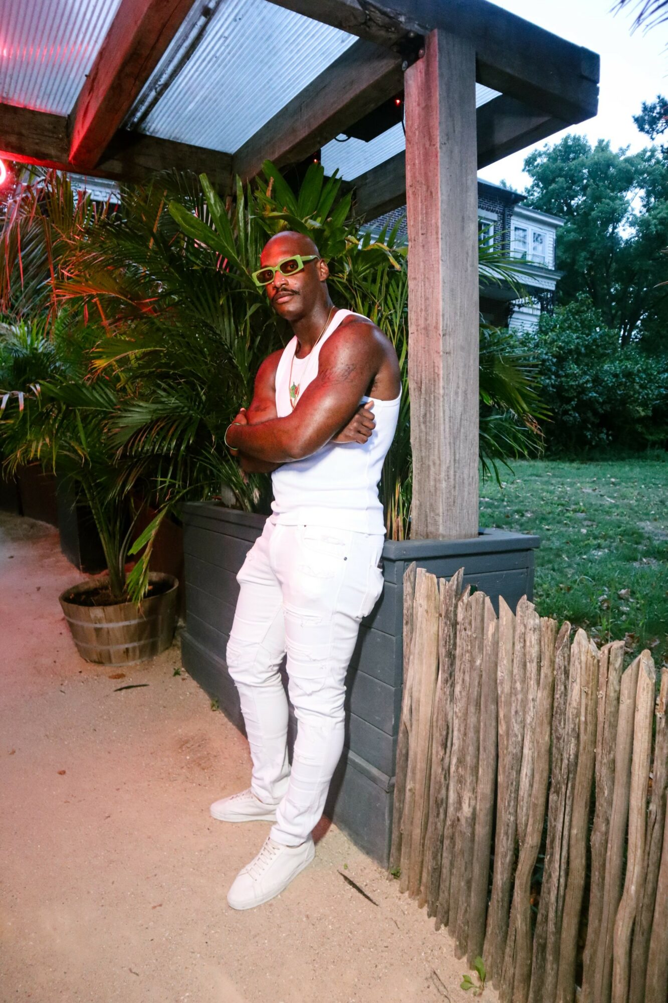 Person in white outfit and sunglasses standing outdoors near plants and a wooden fence.
