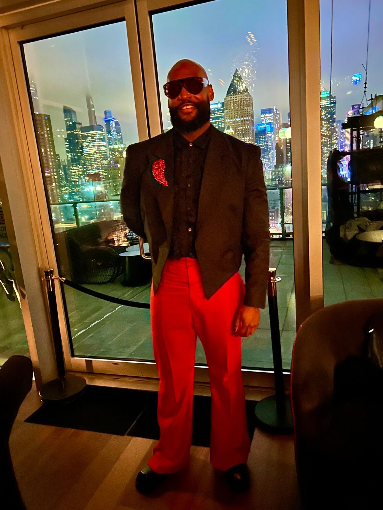 Man in black blazer and red pants standing indoors with city skyline at night visible through large windows behind him.
