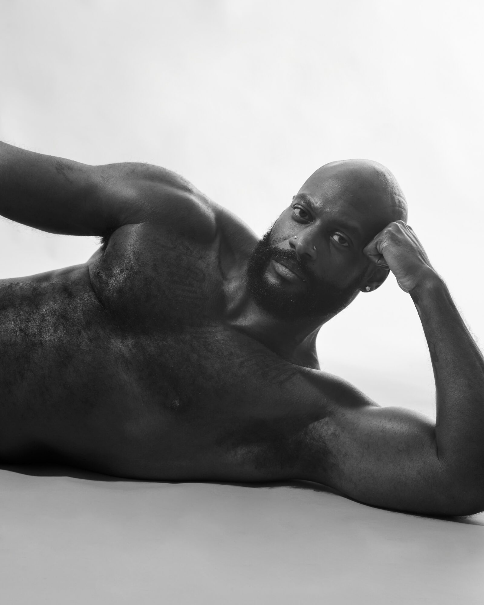 A shirtless man lies on his side, supporting his head with his hand, looking at the camera, in black and white.