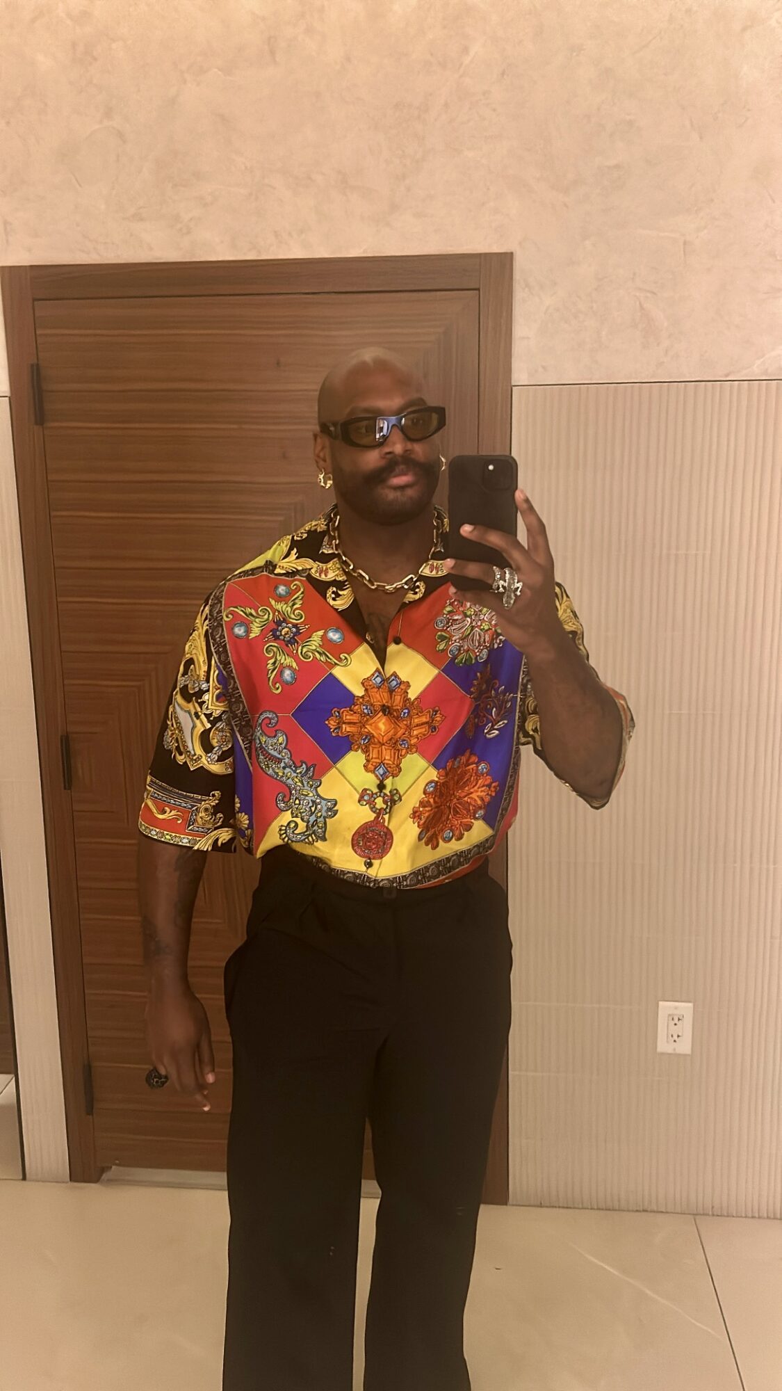 Person taking a mirror selfie, wearing sunglasses and a colorful patterned shirt, standing in front of a wooden door.