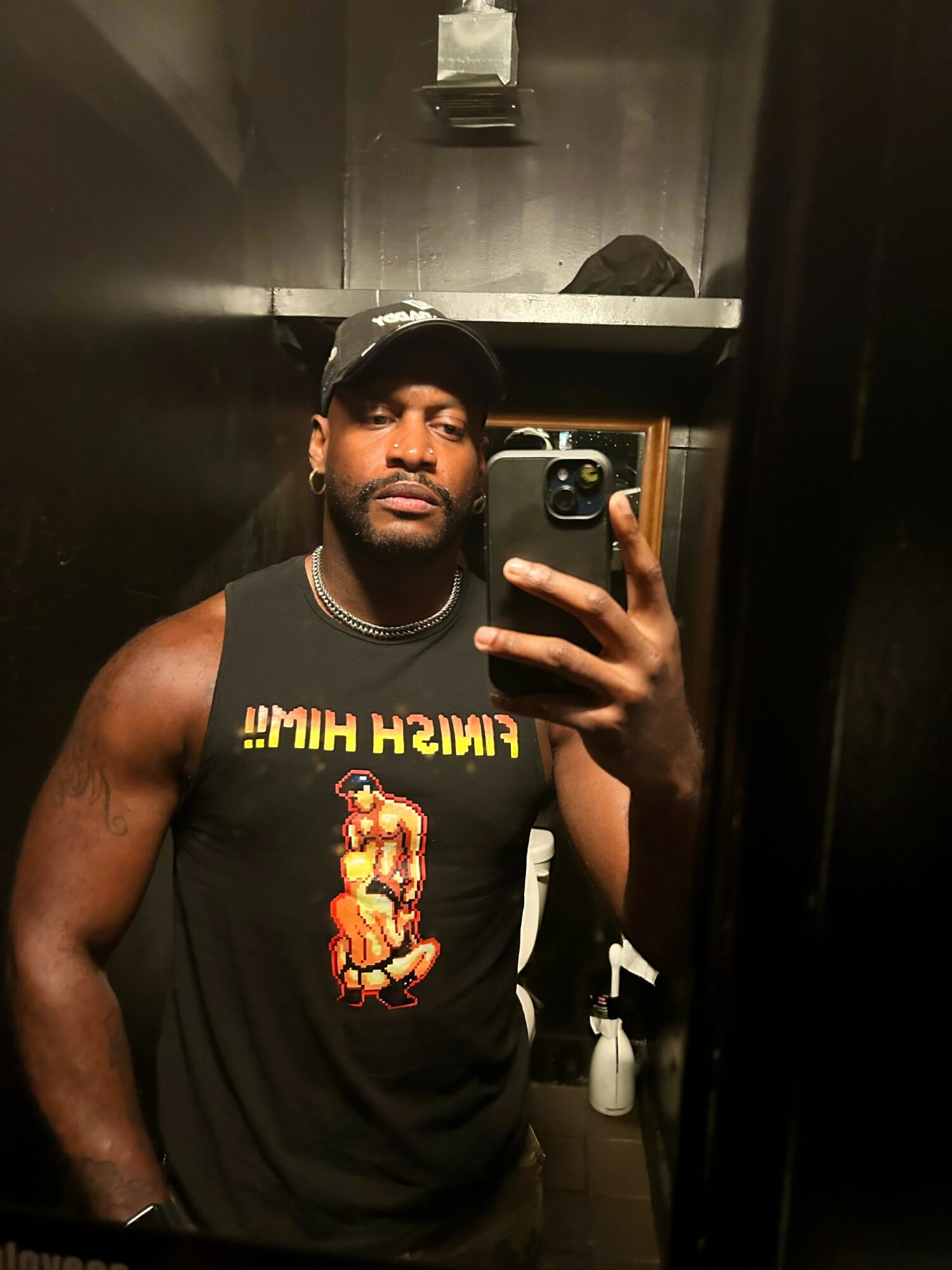Man taking a mirror selfie in a dark room, wearing a black tank top with a graphic and a cap.