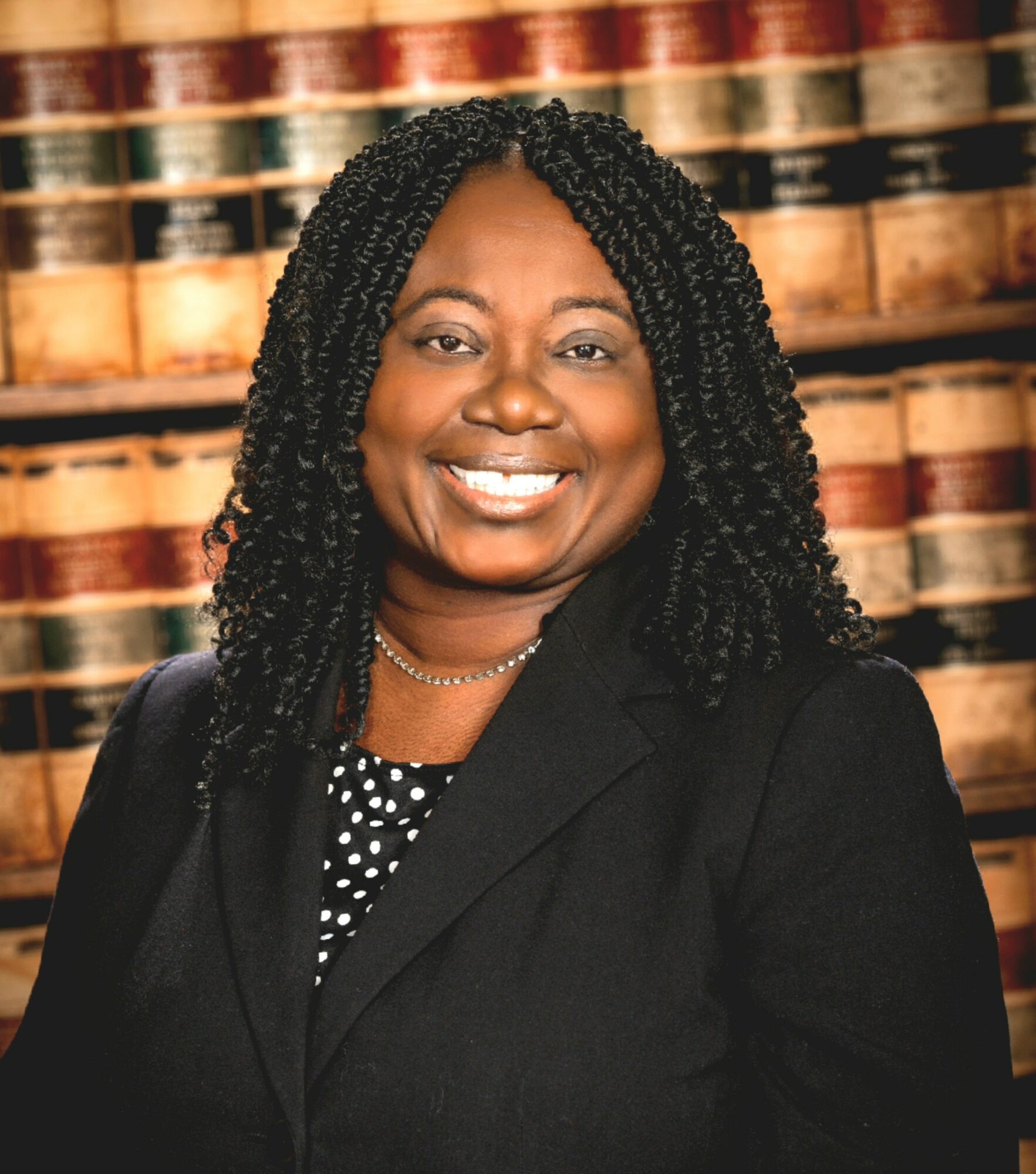 Smiling woman with curly hair in a black blazer, background of stacked books, wearing a polka dot top.
