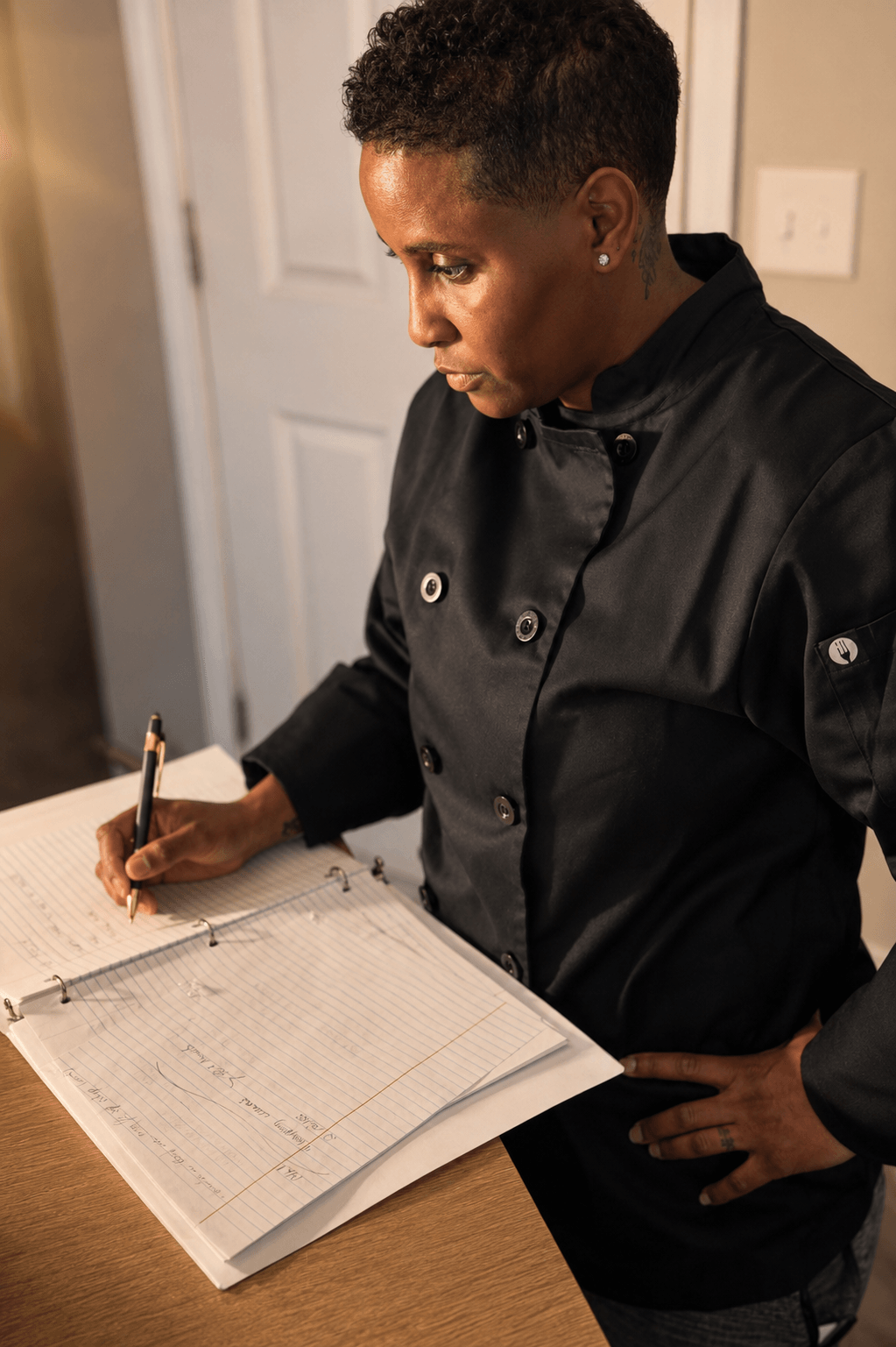 Woman with short curly hair writing in a notebook on a table, wearing a black jacket, indoors.