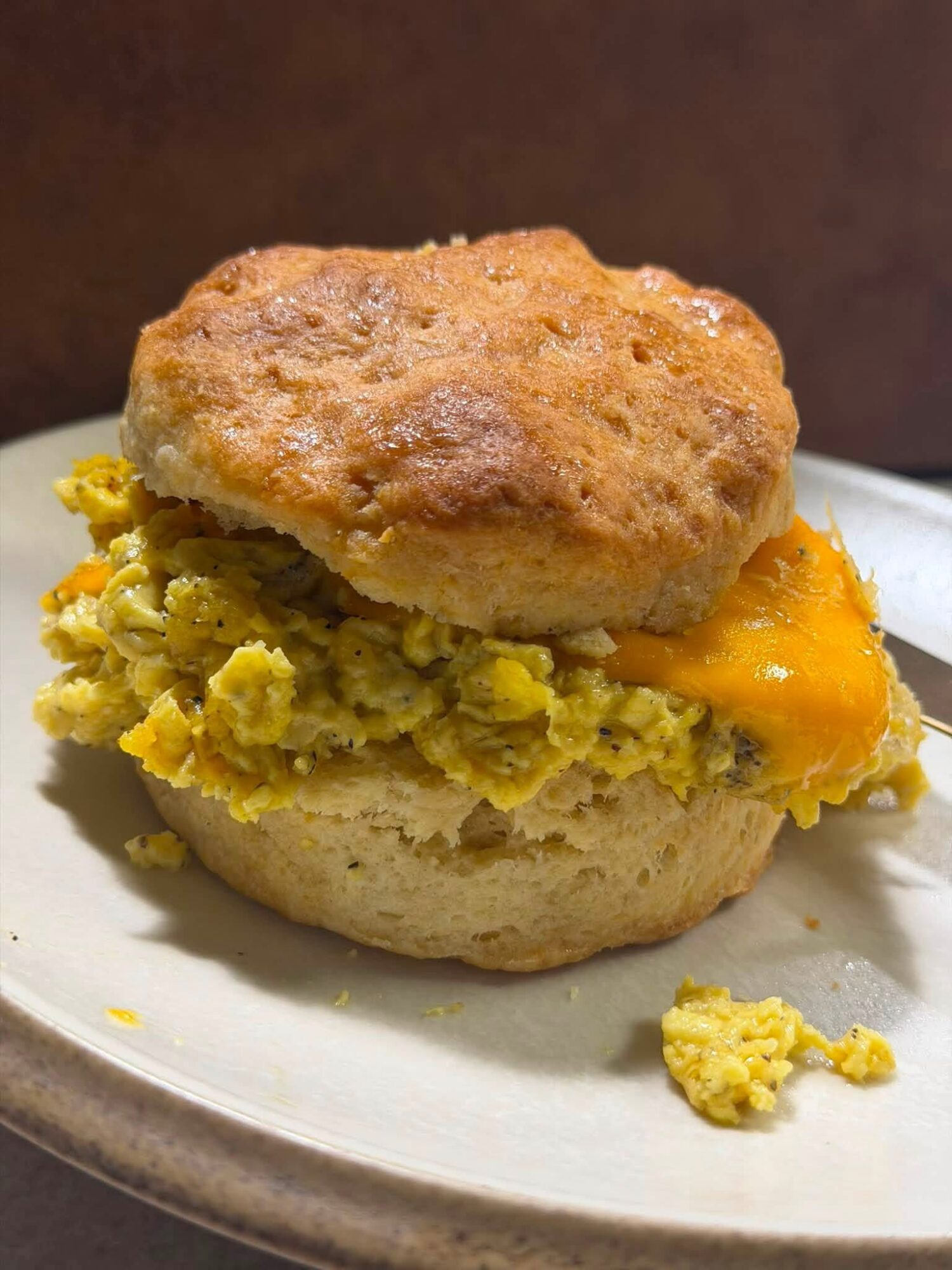 A biscuit sandwich with scrambled eggs and melted cheese inside, topped with a sausage patty, on a plate.