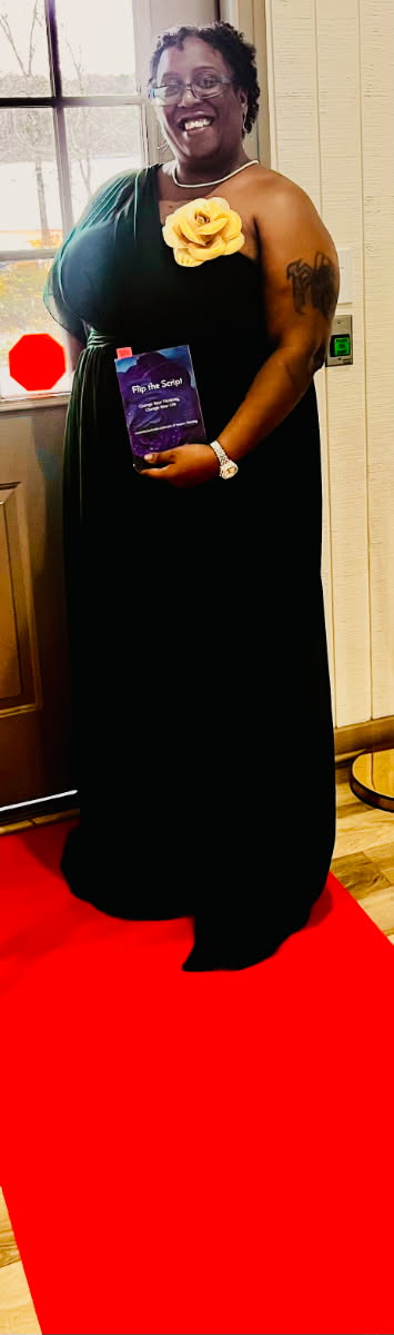 Woman in black dress holding a book, smiling, standing on a red carpet, with a yellow flower on her shoulder.