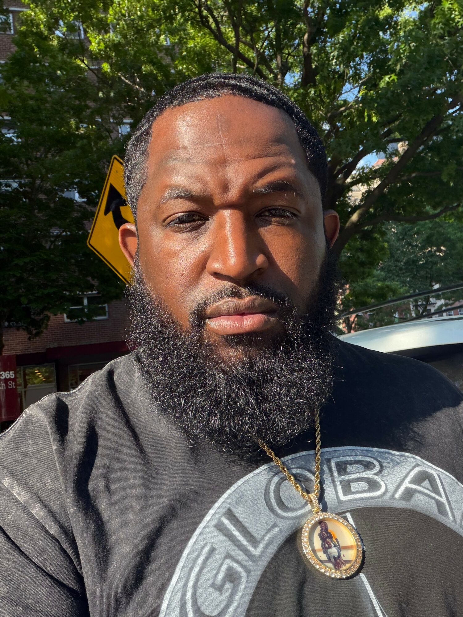 Close-up of a man with a beard and mustache outdoors, trees and a yellow road sign in background, wearing a black shirt and gold necklace.