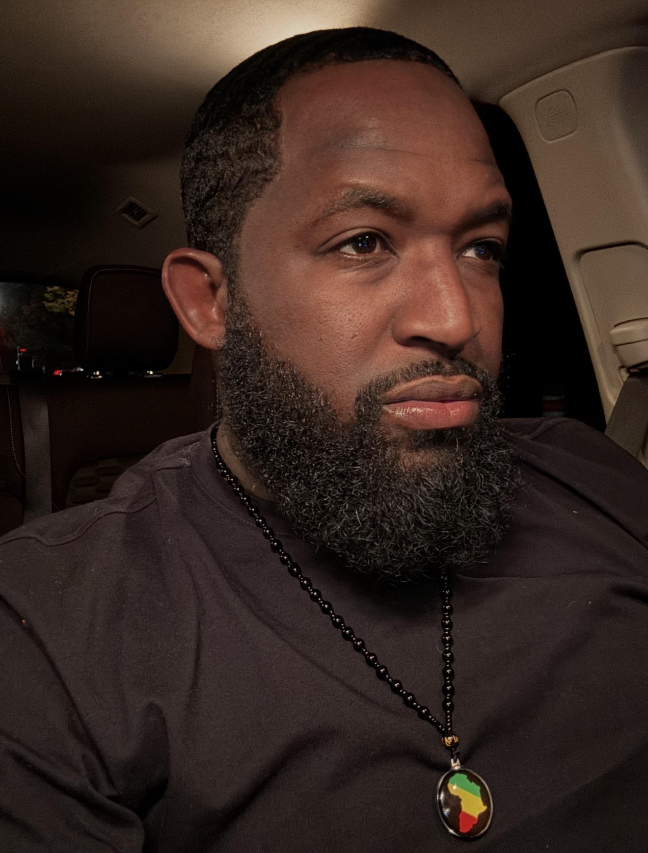 Man with beard and short hair wearing a black shirt and a colorful beaded necklace inside a vehicle.